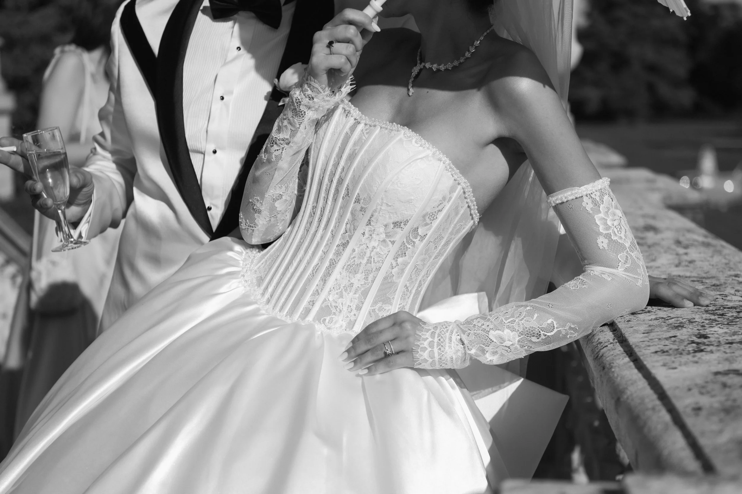 Bride in a lace wedding gown with lace gloves and a veil, wedding rings, and a necklace, leaning on a stone railing, with a groom in a tuxedo holding a glass of champagne and a cigarette in the background.