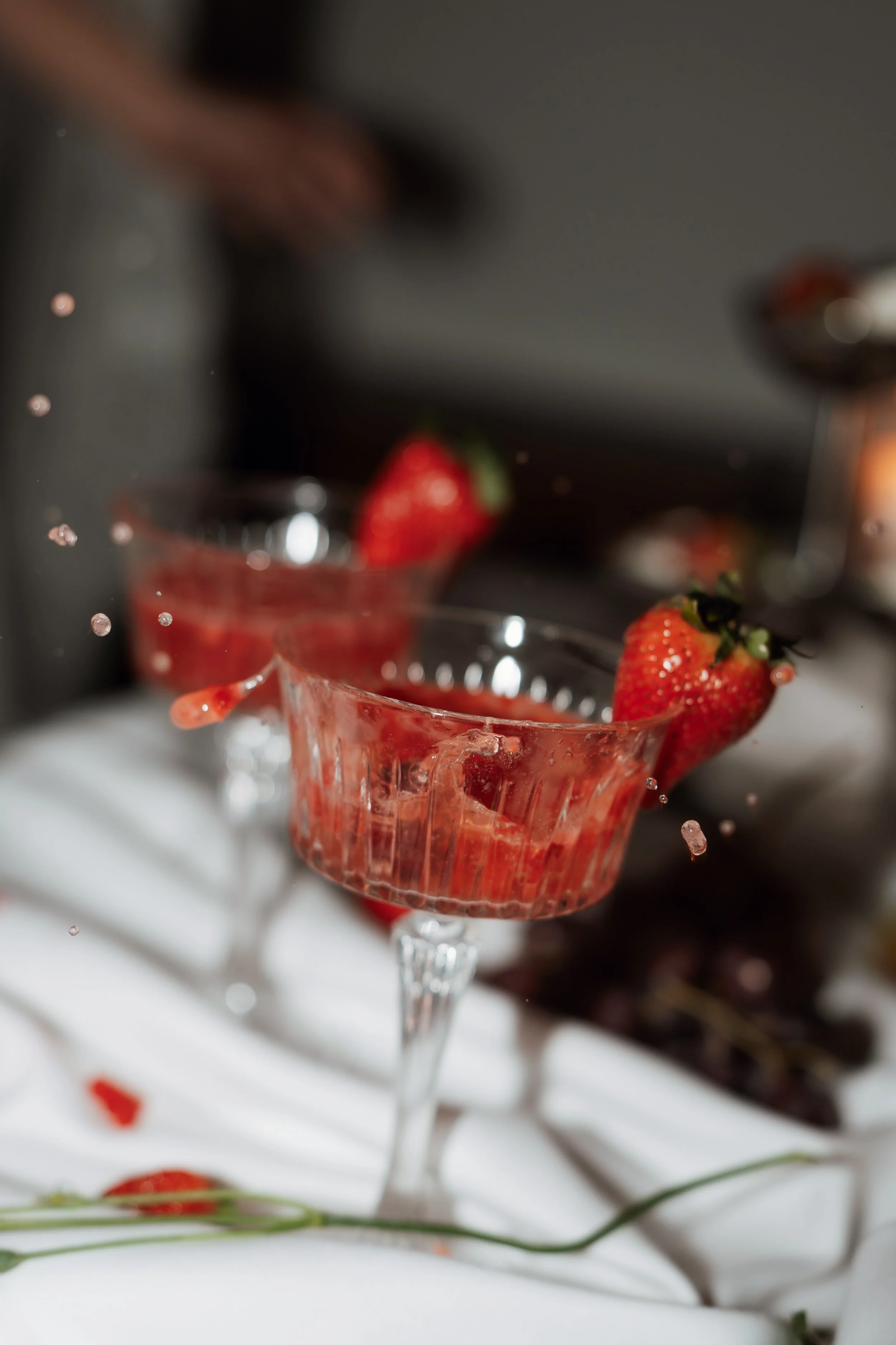 Two pink cocktails with strawberries garnishing the rim of each glass, with droplets of liquid capturing mid-air, set on a white surface with scattered strawberries and dark background.