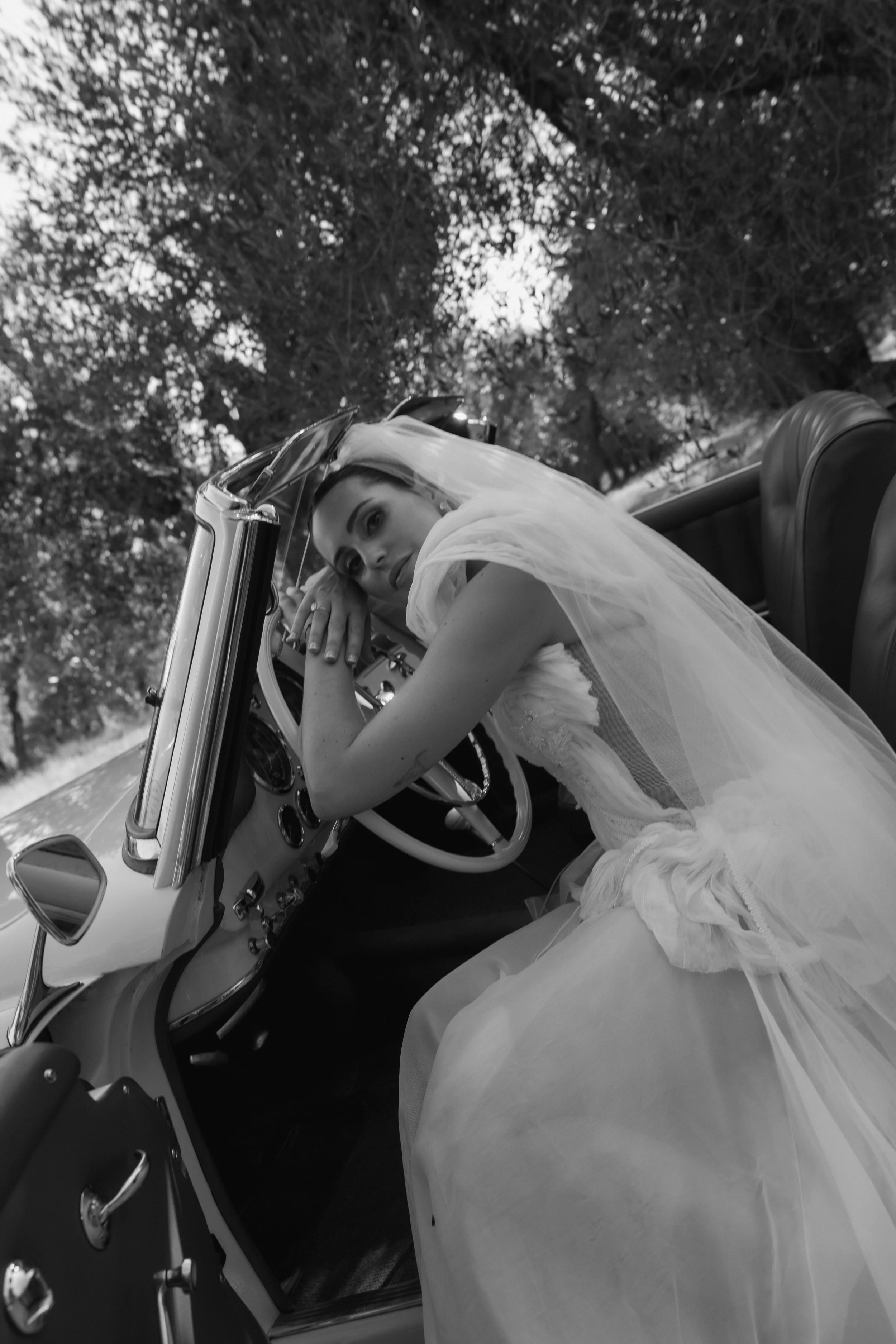 A woman in a wedding dress leaning on the steering wheel of a vintage car, with trees in the background.