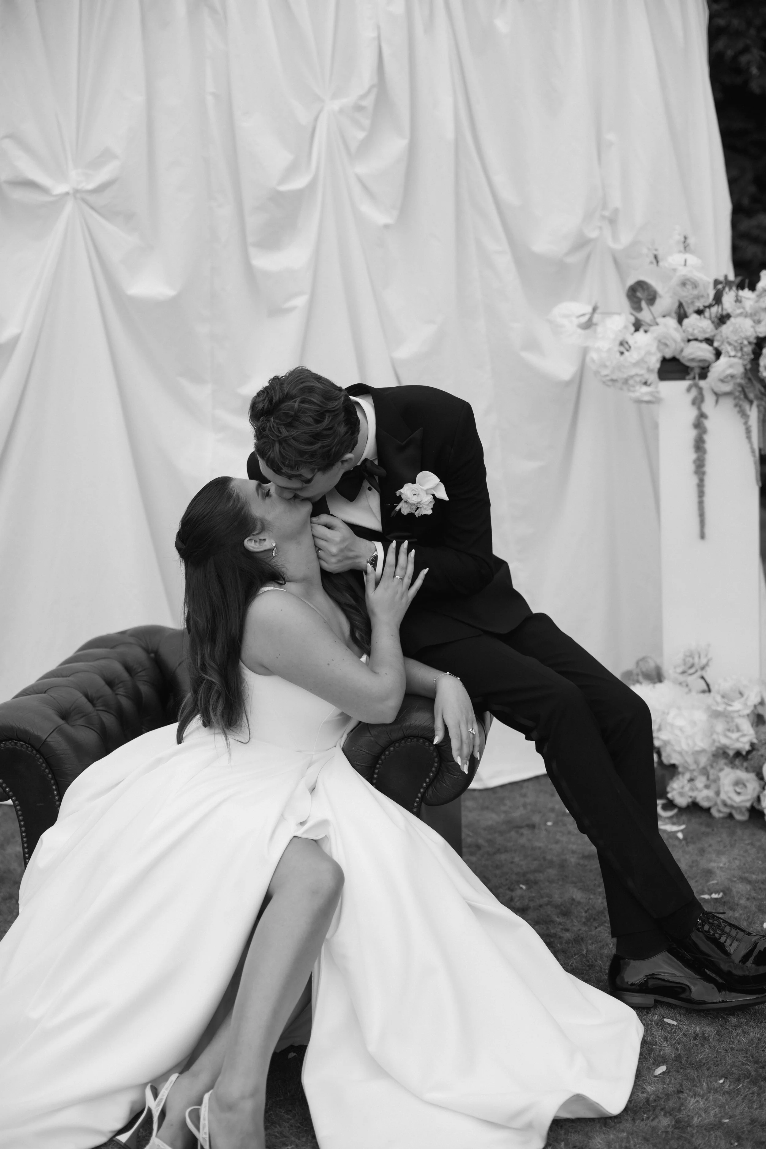 A black and white photo of a bride and groom sharing a kiss during their wedding ceremony, with the bride sitting on a vintage-style couch and the groom standing beside her, exchanging a kiss outdoors in front of a decorated backdrop with flowers.