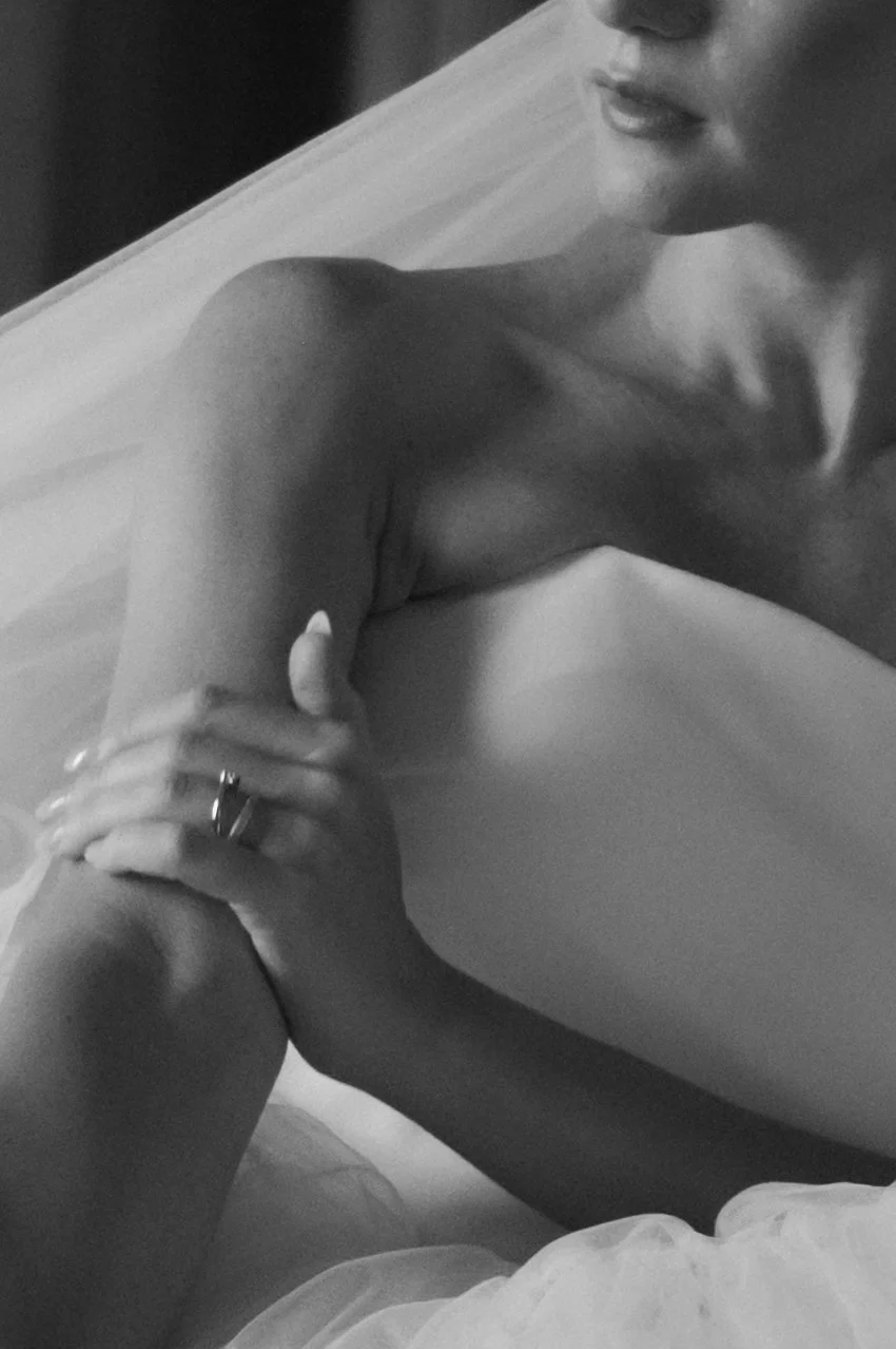 A person with shoulder-length hair and wearing a ring on their finger is sitting on a bed, partially covered by a sheer fabric, in a black and white photograph.