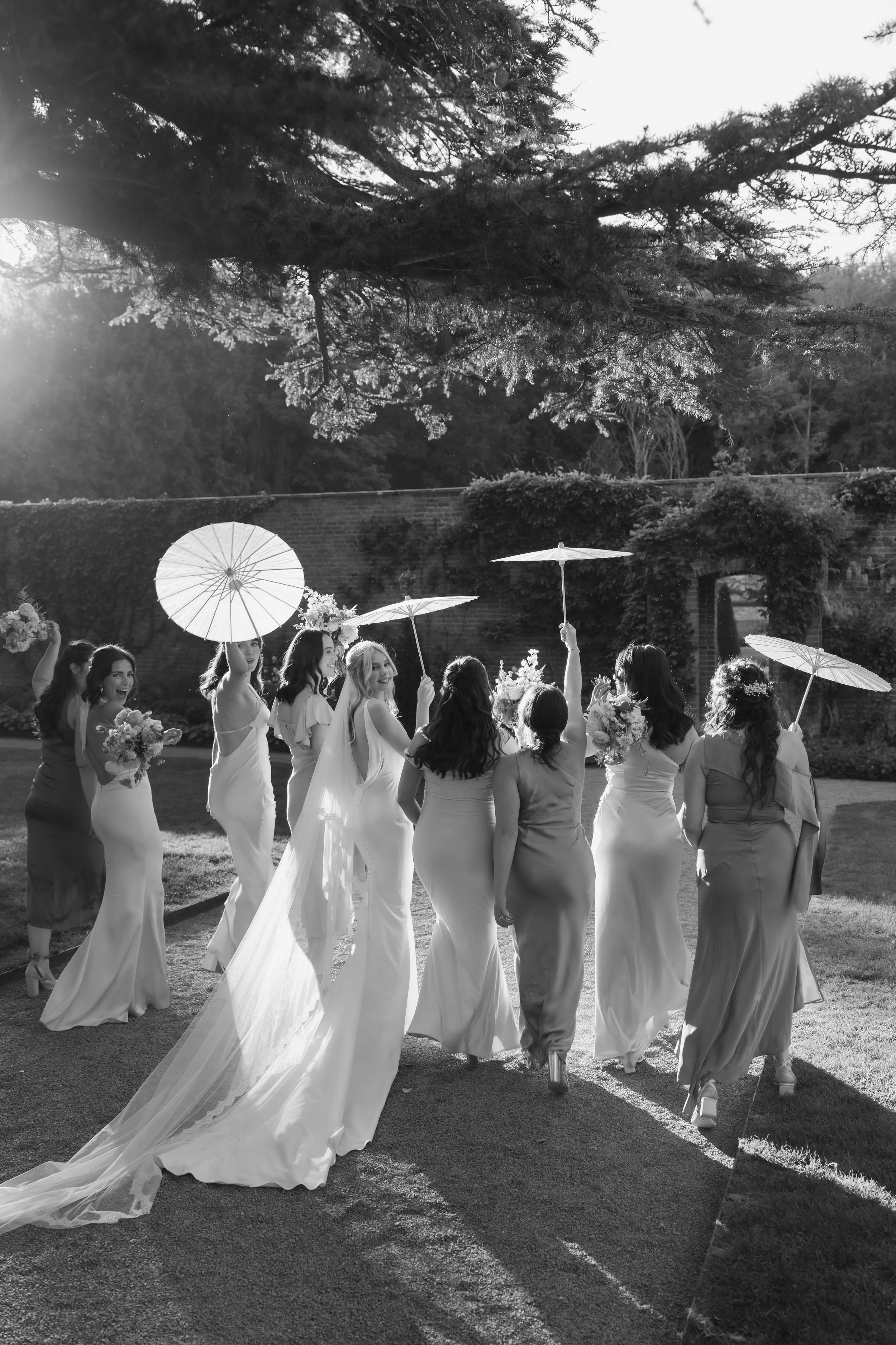 A bride and her bridesmaids walking outdoors, holding umbrellas and bouquets, with sunlight in the background at Garthmyl Hall 