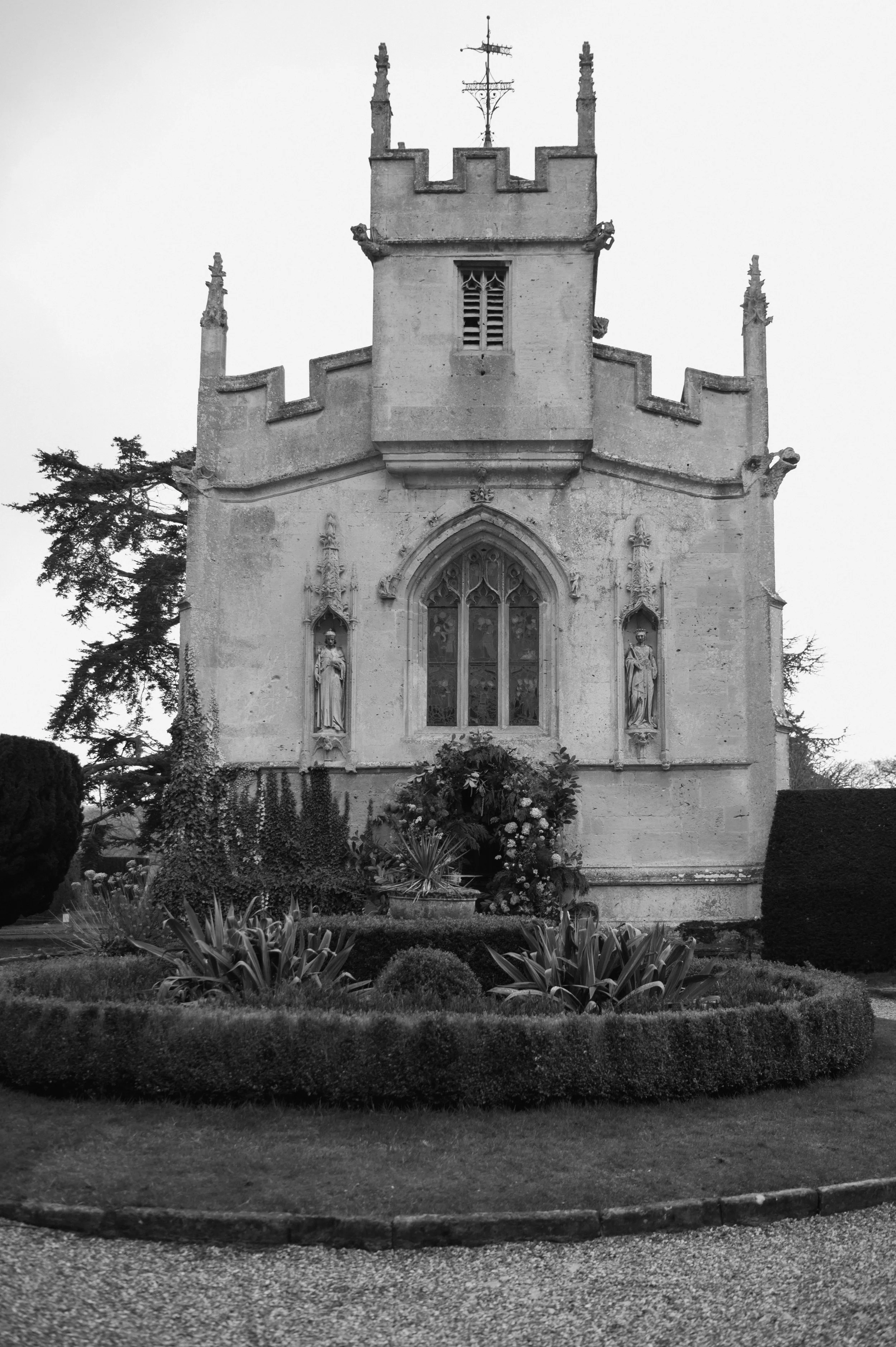 Black and white photo of a historic castle-like building with gothic windows, statues, and a small garden with flowers in front.