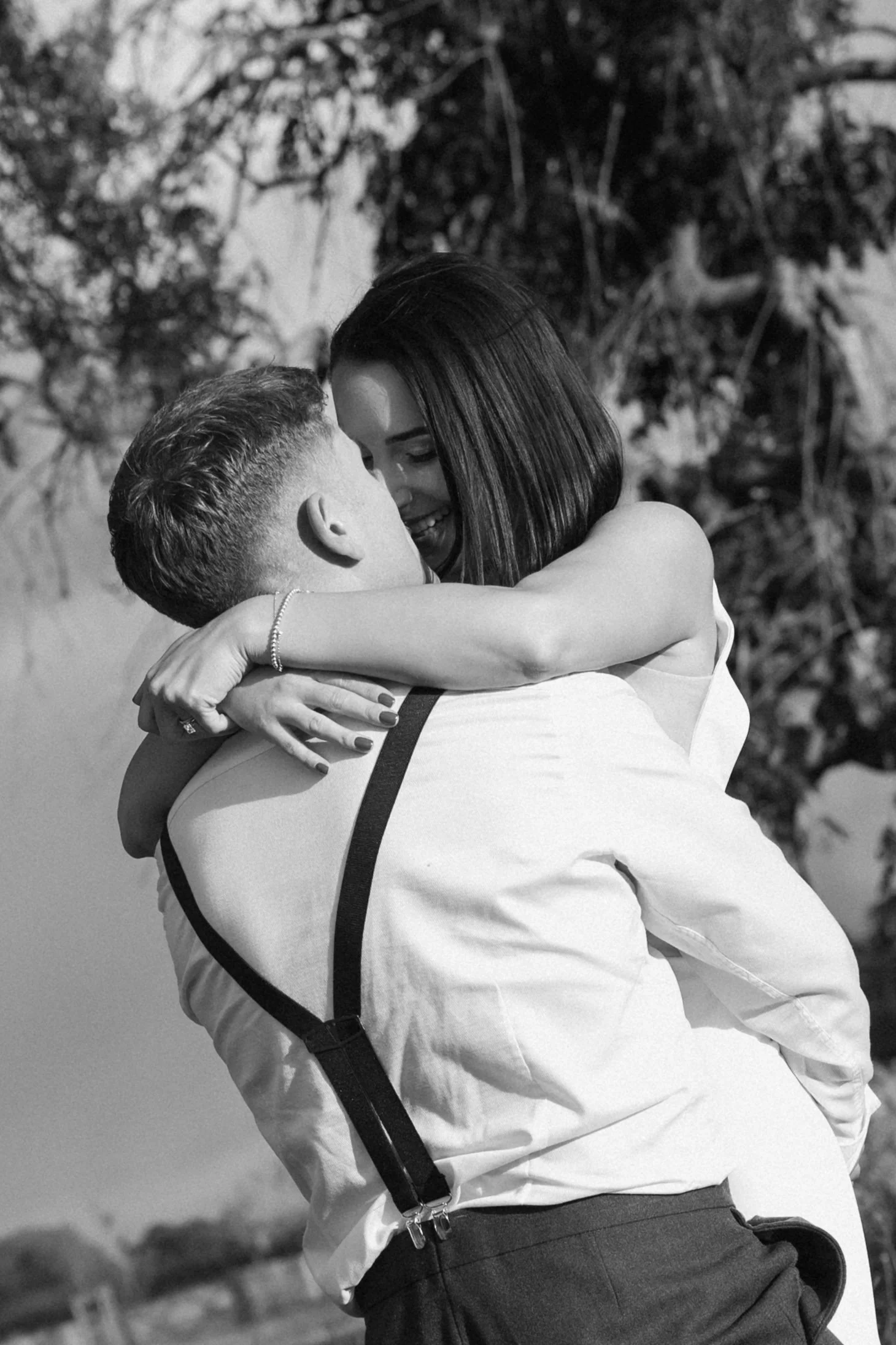 A couple embracing and kissing outdoors, black and white photo, woman with dark hair and man with short hair in formal attire.