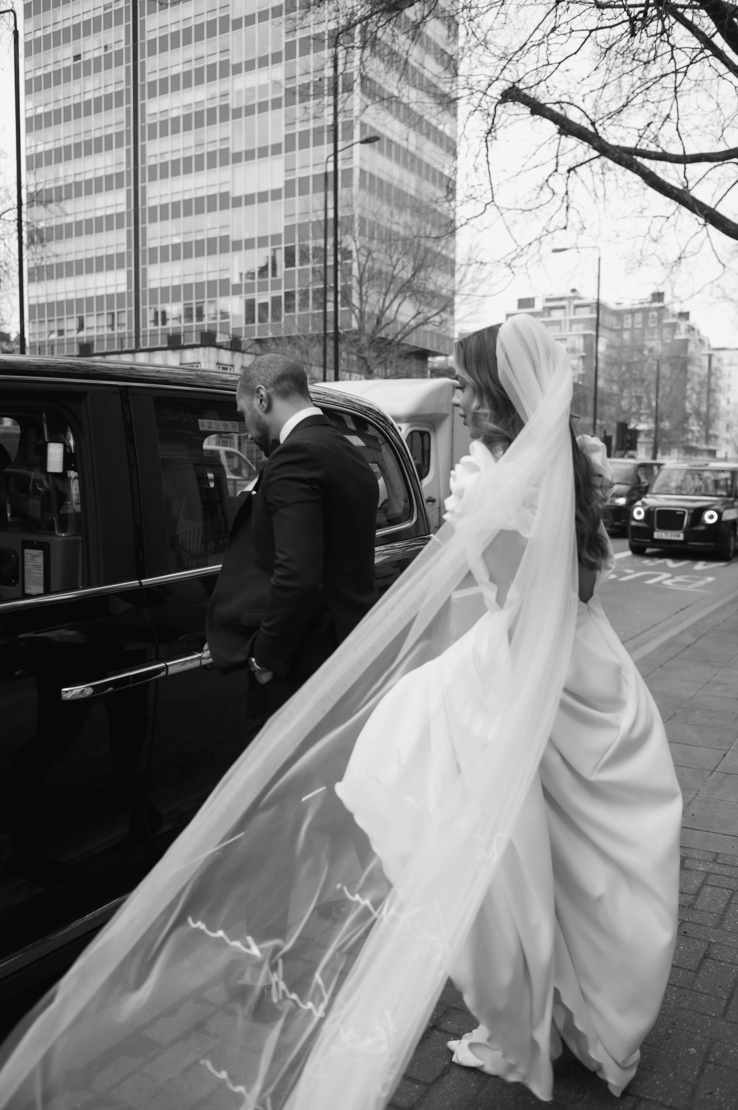 A bride in a wedding dress and veil standing outside next to a black taxi, with a groom in a suit inside the taxi, on a city street with tall buildings and leafless trees.