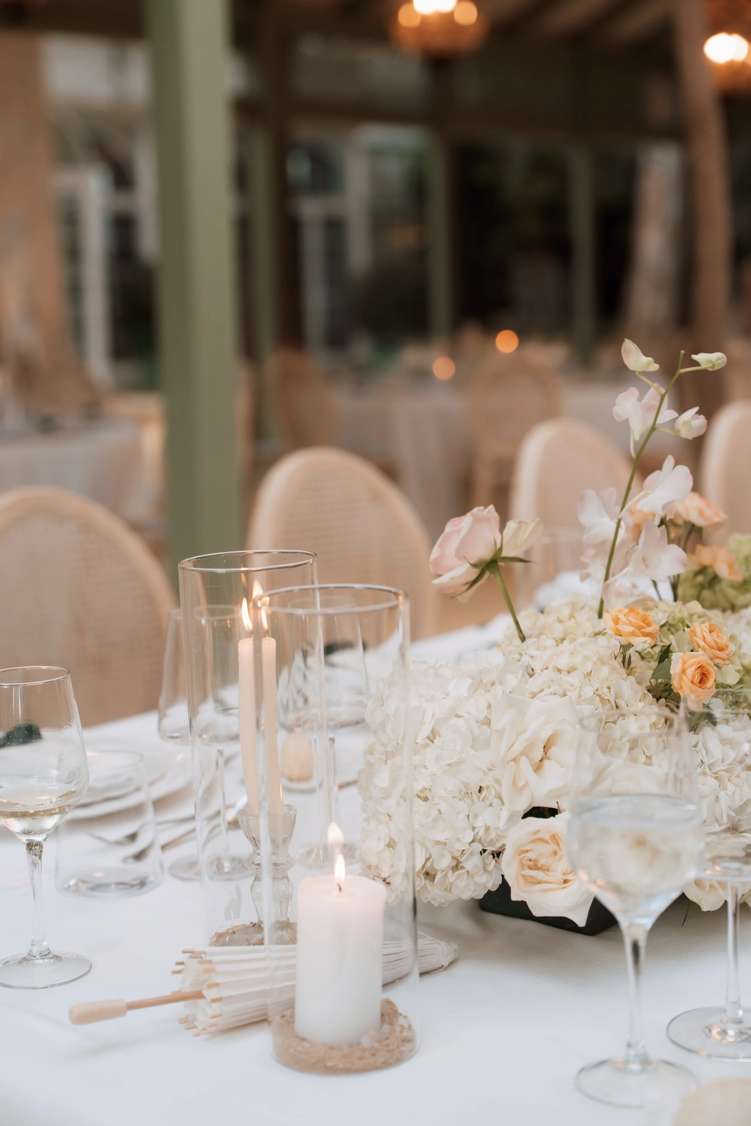 A decorated table set for an event, featuring white hydrangeas and peach roses in a centerpiece, surrounded by glass candles and wine glasses, with chairs in the background.