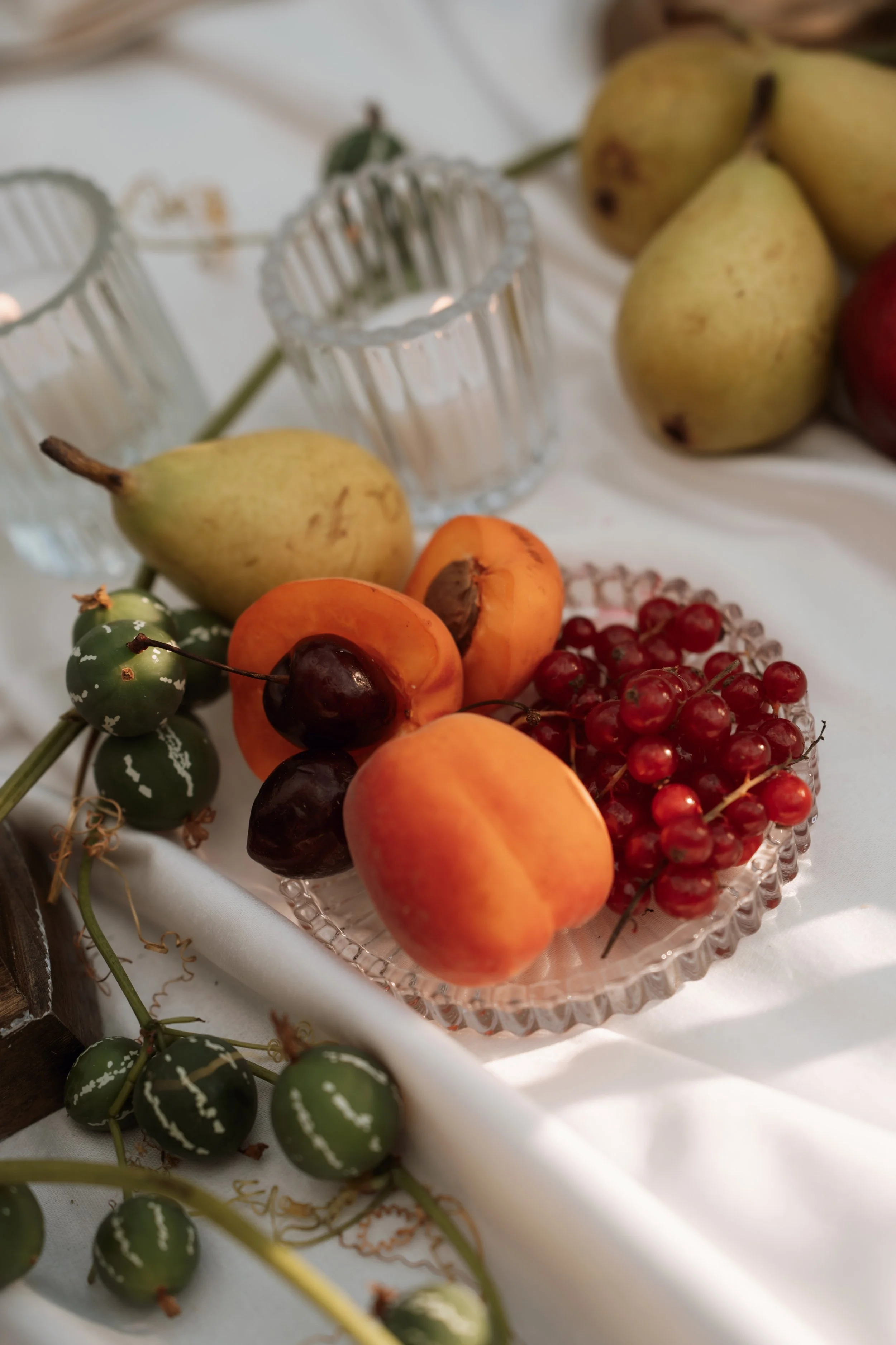 Arrangement of various fruits on a white tablecloth, including green pears, apricots, cherries, and red currants, with glass candle holders nearby.