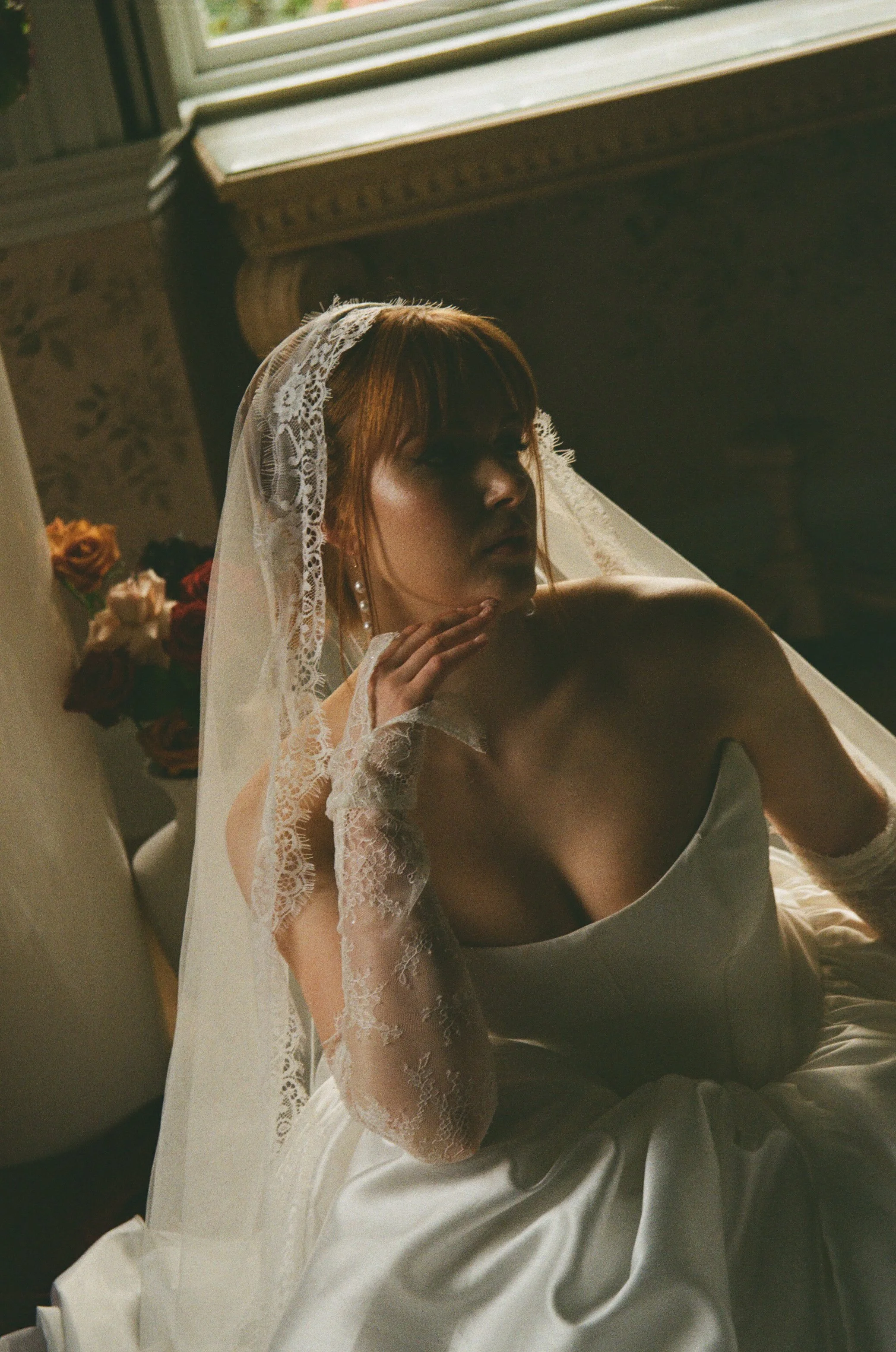 Bride in lace gloves and veil sitting by window with flowers, natural light taken on 35mm film photography