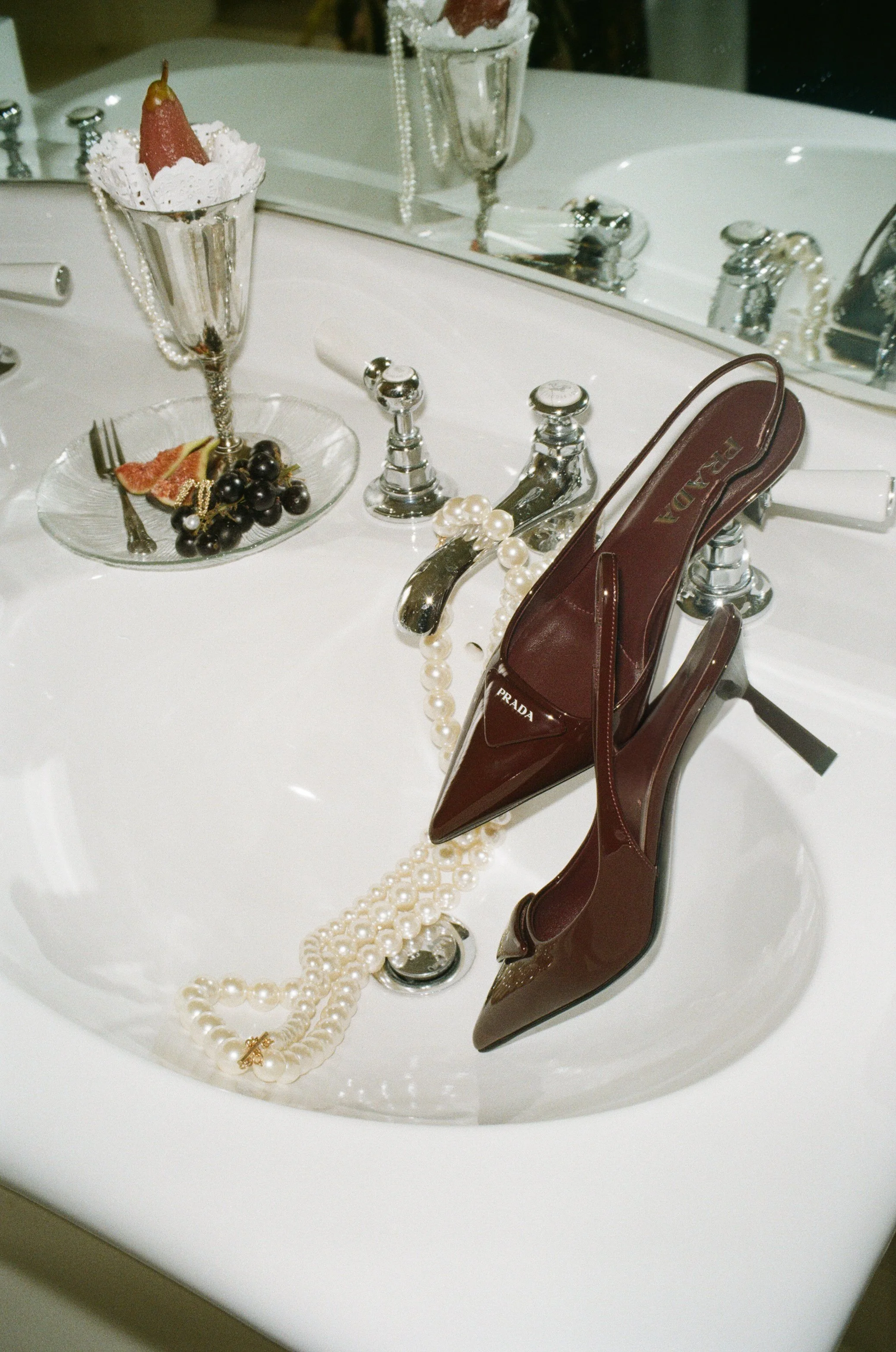 Bathroom sink with pearl necklace, burgundy Prada heels, figs, grapes, and glass at Iscoyd park 