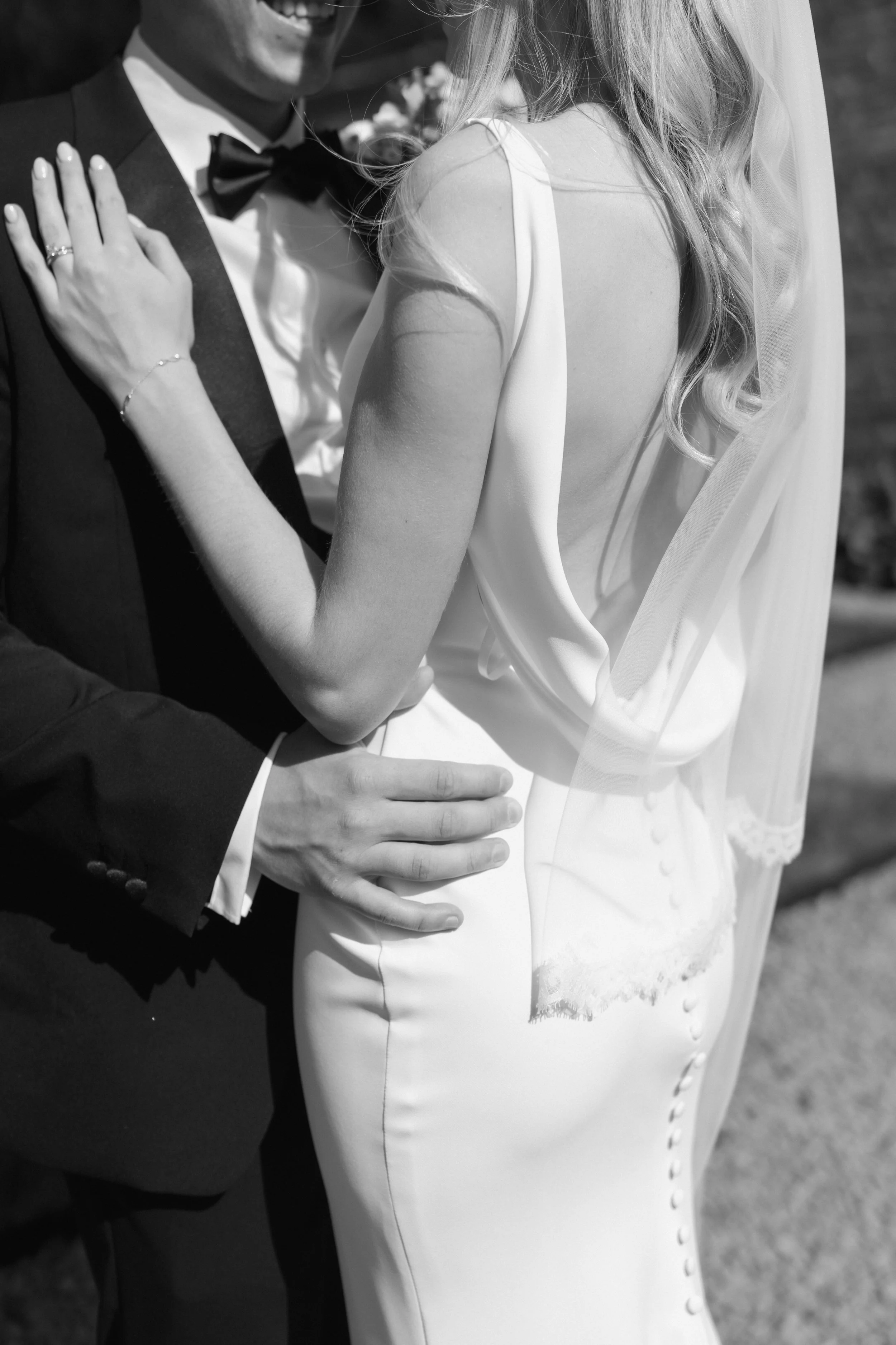 Close-up of a bride and groom on their wedding day, showing the bride's back in a white dress with buttons and a veil, and the groom in a tuxedo with a bow tie.