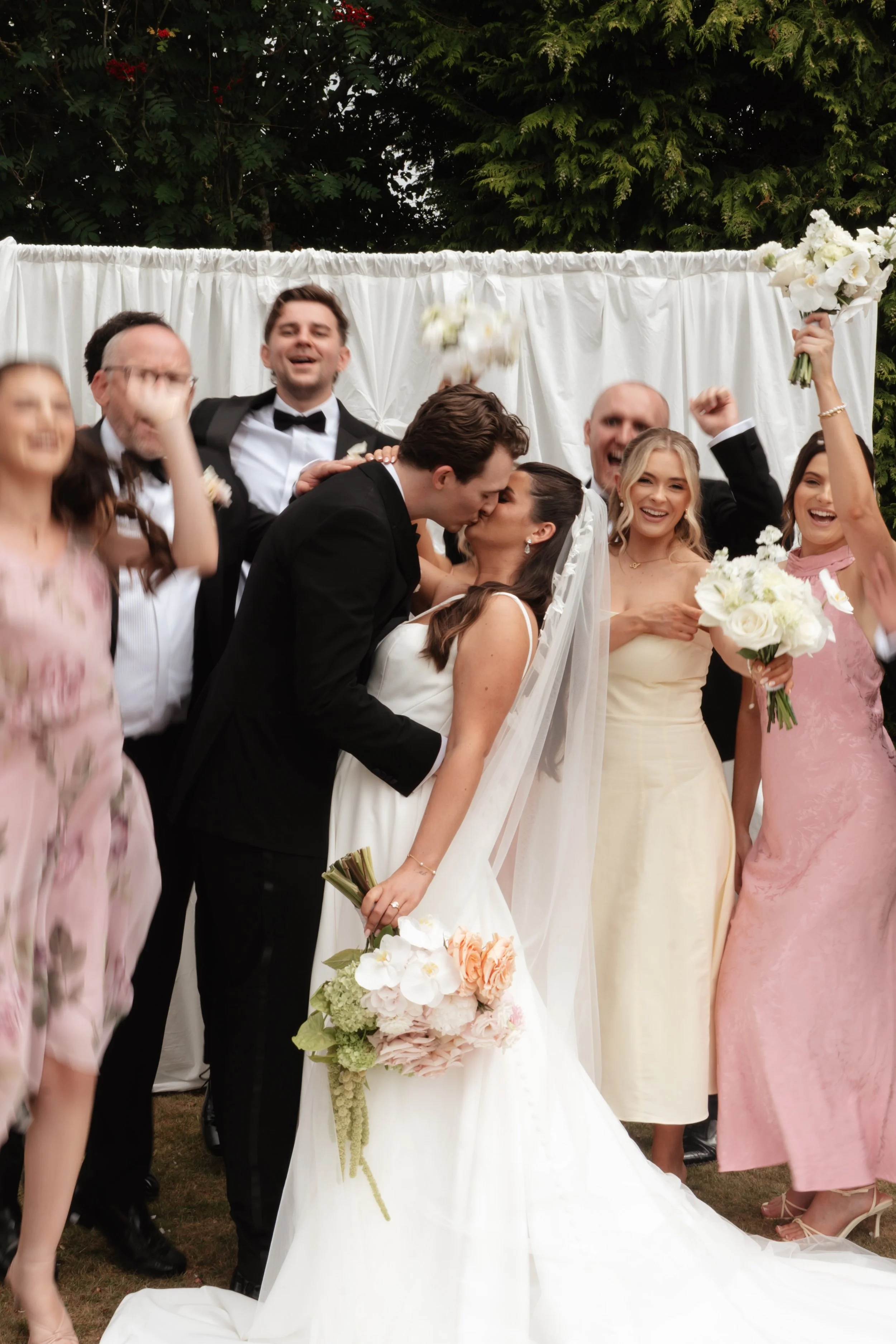 A wedding celebration with the bride and groom sharing a kiss, surrounded by friends and family, some raising bouquets and smiling cheerfully.