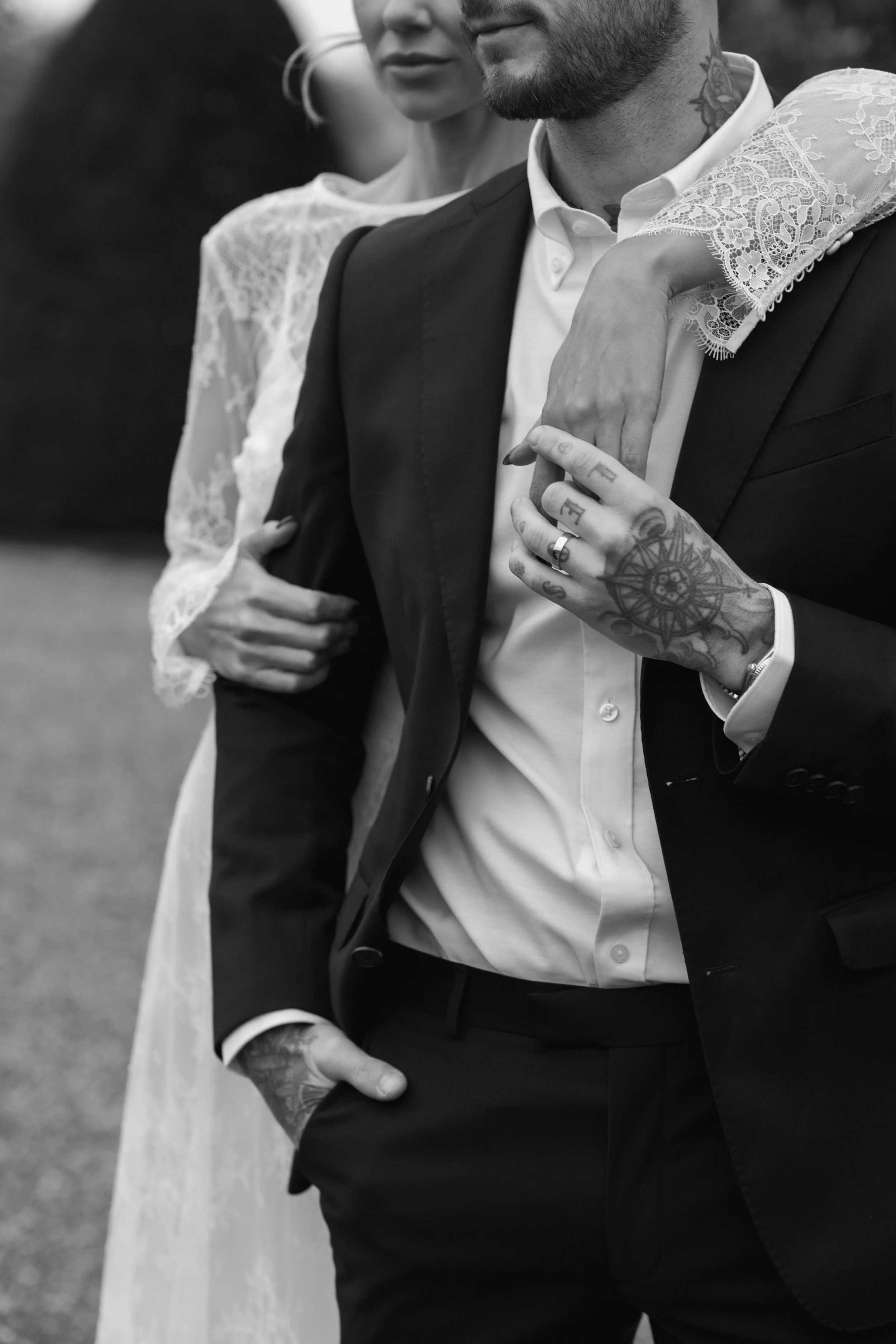 Black and white photo of a couple, with the woman resting her arm on the man's shoulder. The man is dressed in a suit with tattoos visible on his hand and neck. The woman is wearing a lace dress.