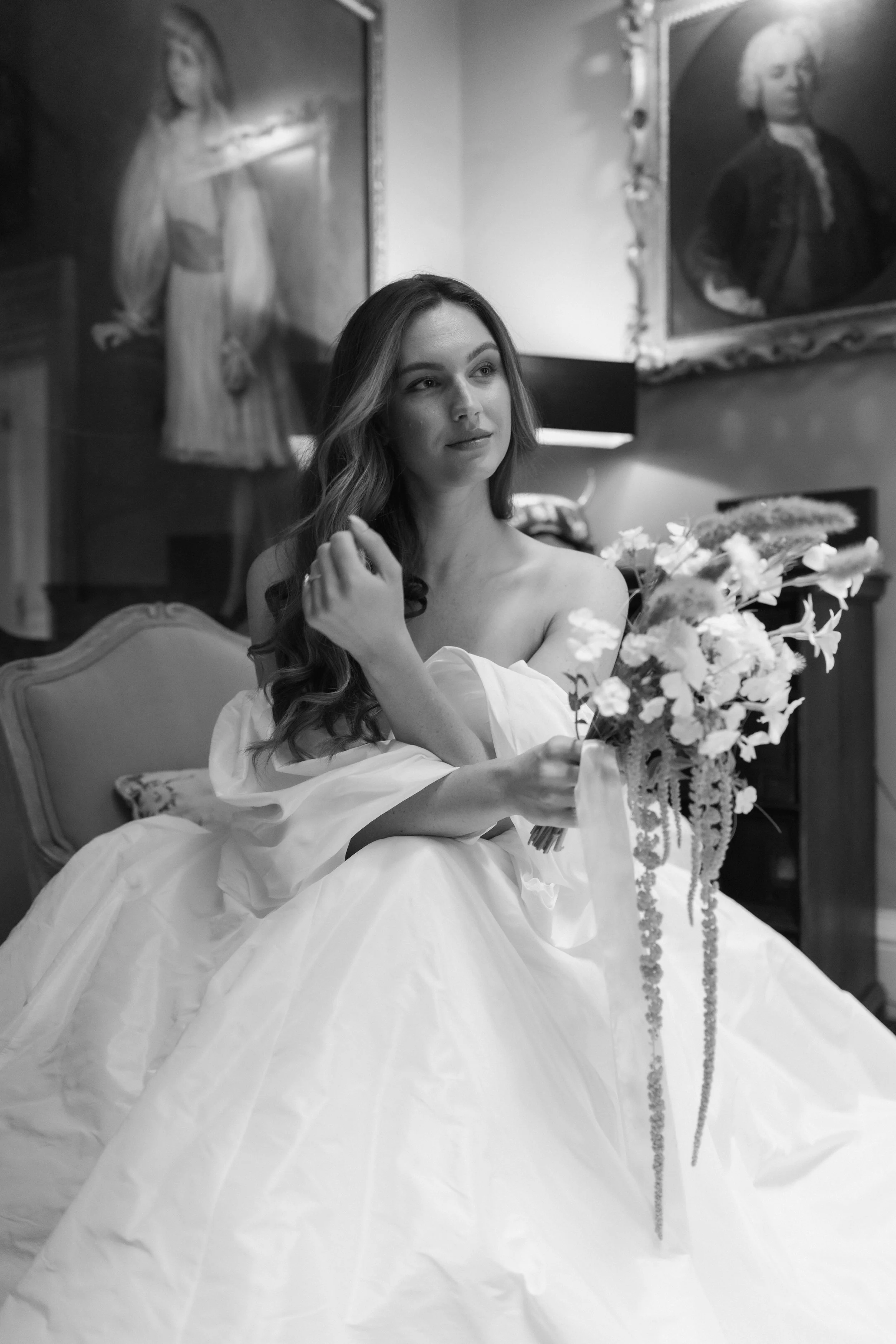 A woman in a wedding dress holding a bouquet of flowers, sitting in a room with two large framed portraits on the wall behind her.