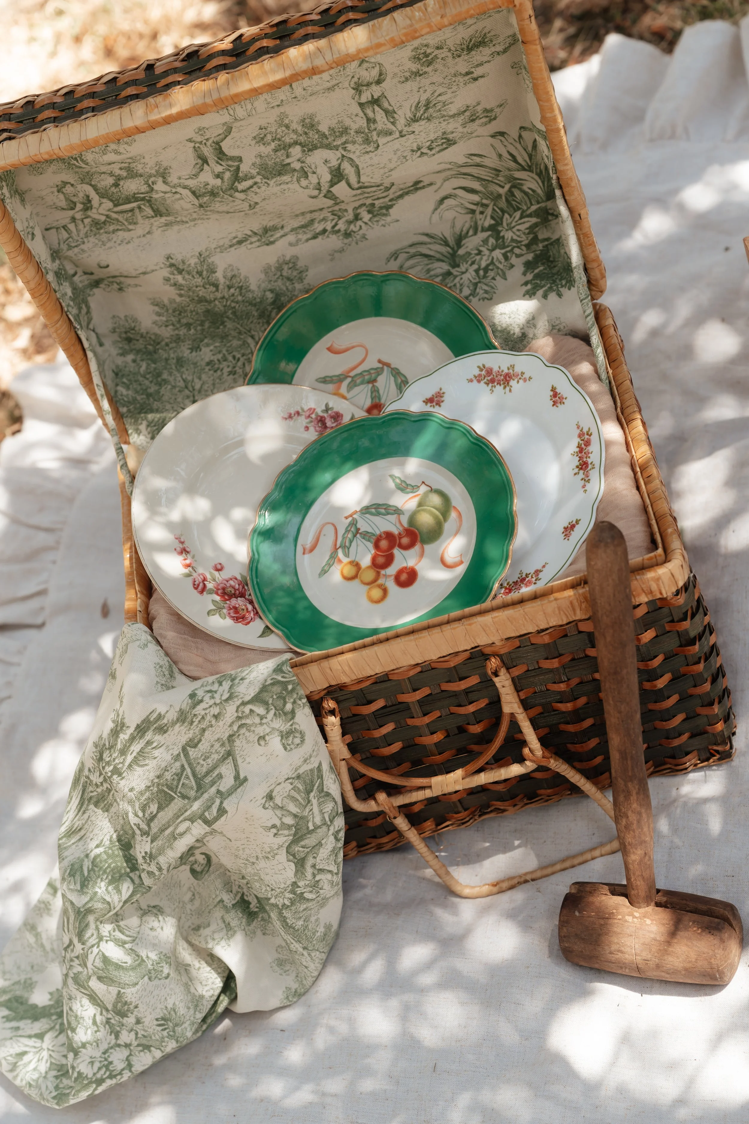 A basket of decorative plates with fruit images, a cloth with a pastoral scene, and a wooden mallet on a white surface outdoors.