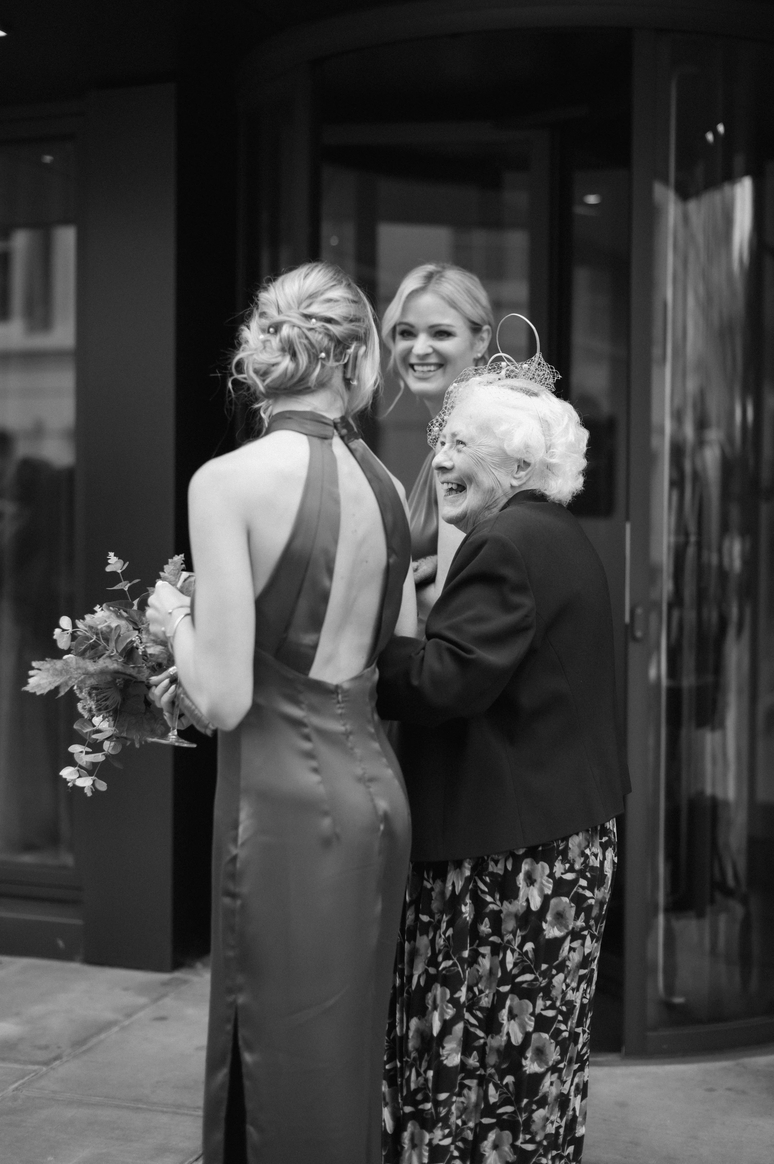 Three women sharing a joyful moment outside a building, one in formal dress holding flowers, an older woman smiling at her, and another woman smiling in the background.