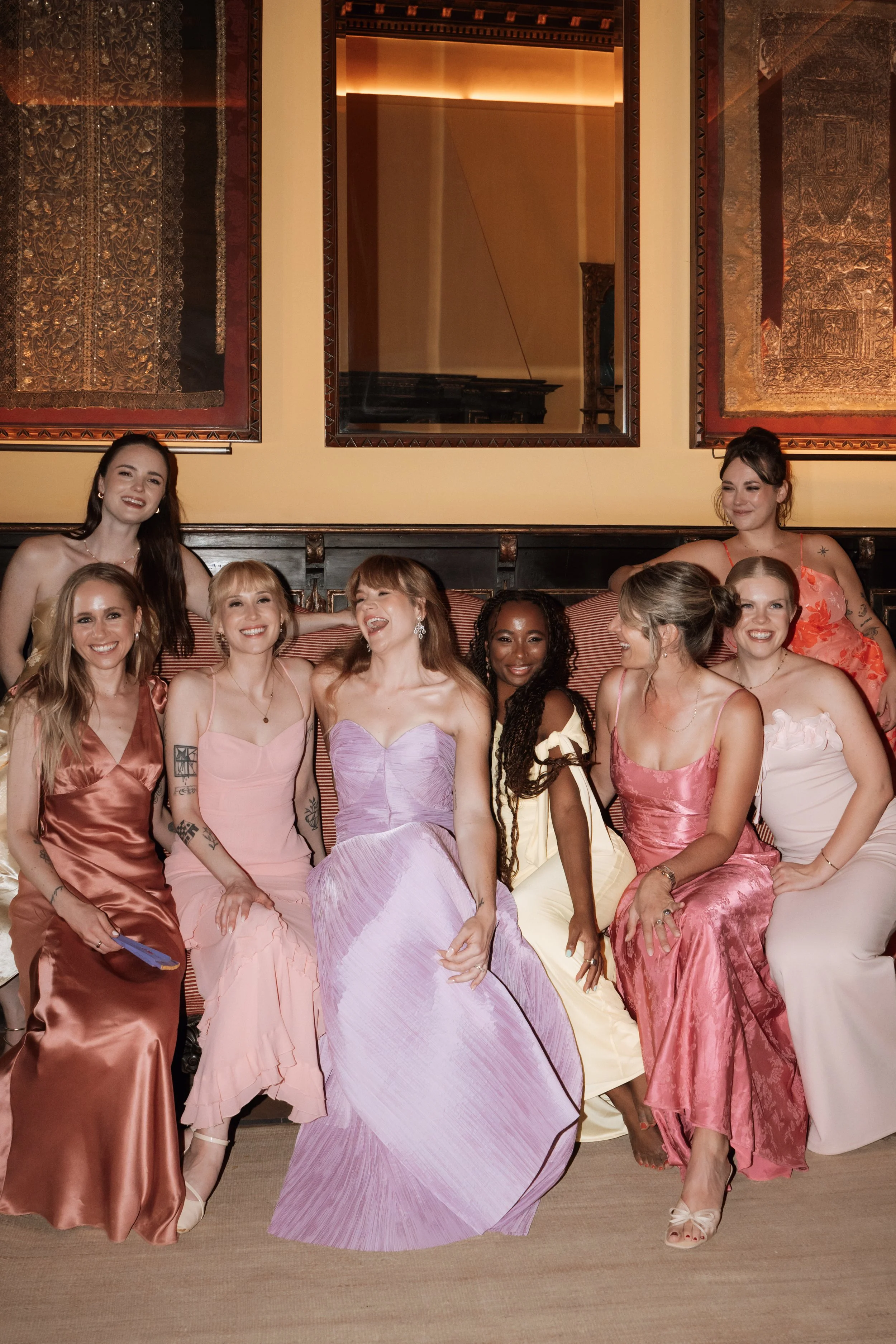 Group of women in colorful dresses sitting on a couch and smiling for a photo in an elegant room with framed artwork on the wall.