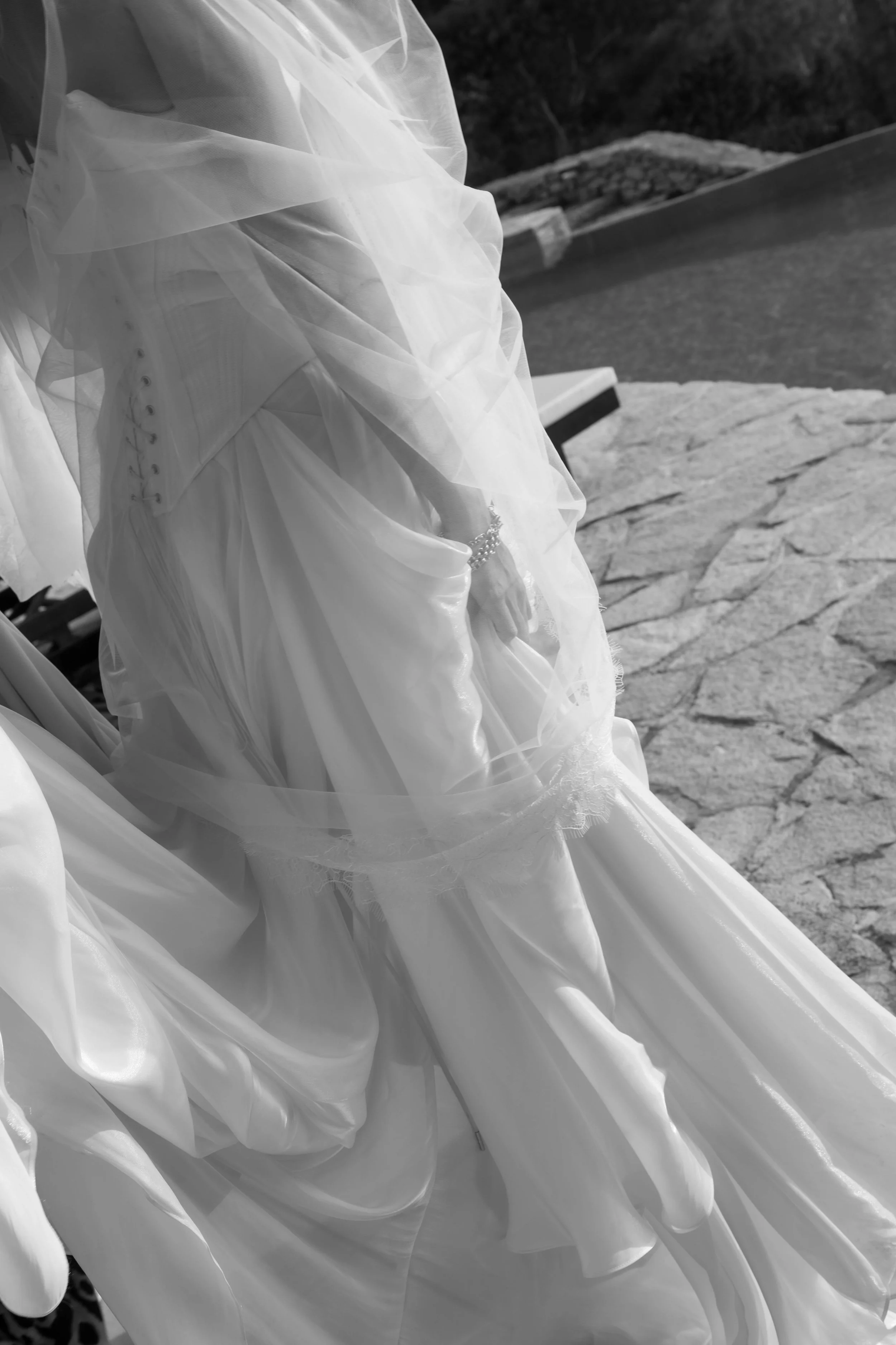 A bride wearing a wedding dress and veil, sitting outdoors on a stone surface with water and trees in the background.