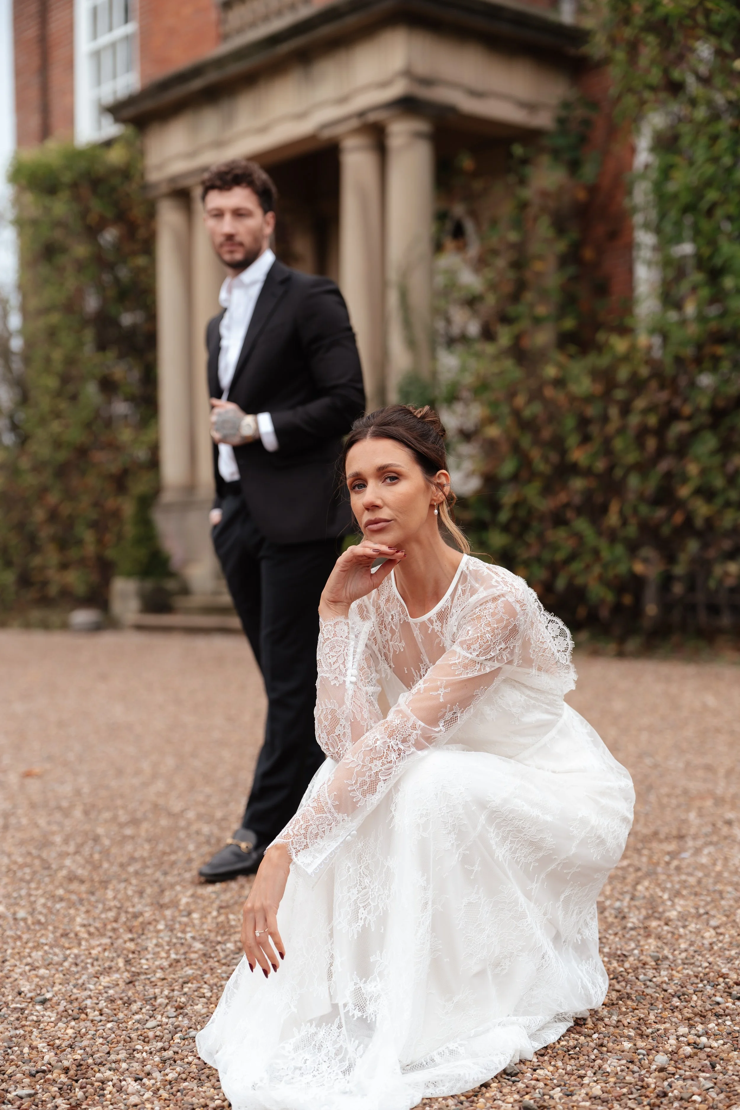 Bride in white lace dress sitting outdoors with groom in black suit behind her in Iscoyd Park 