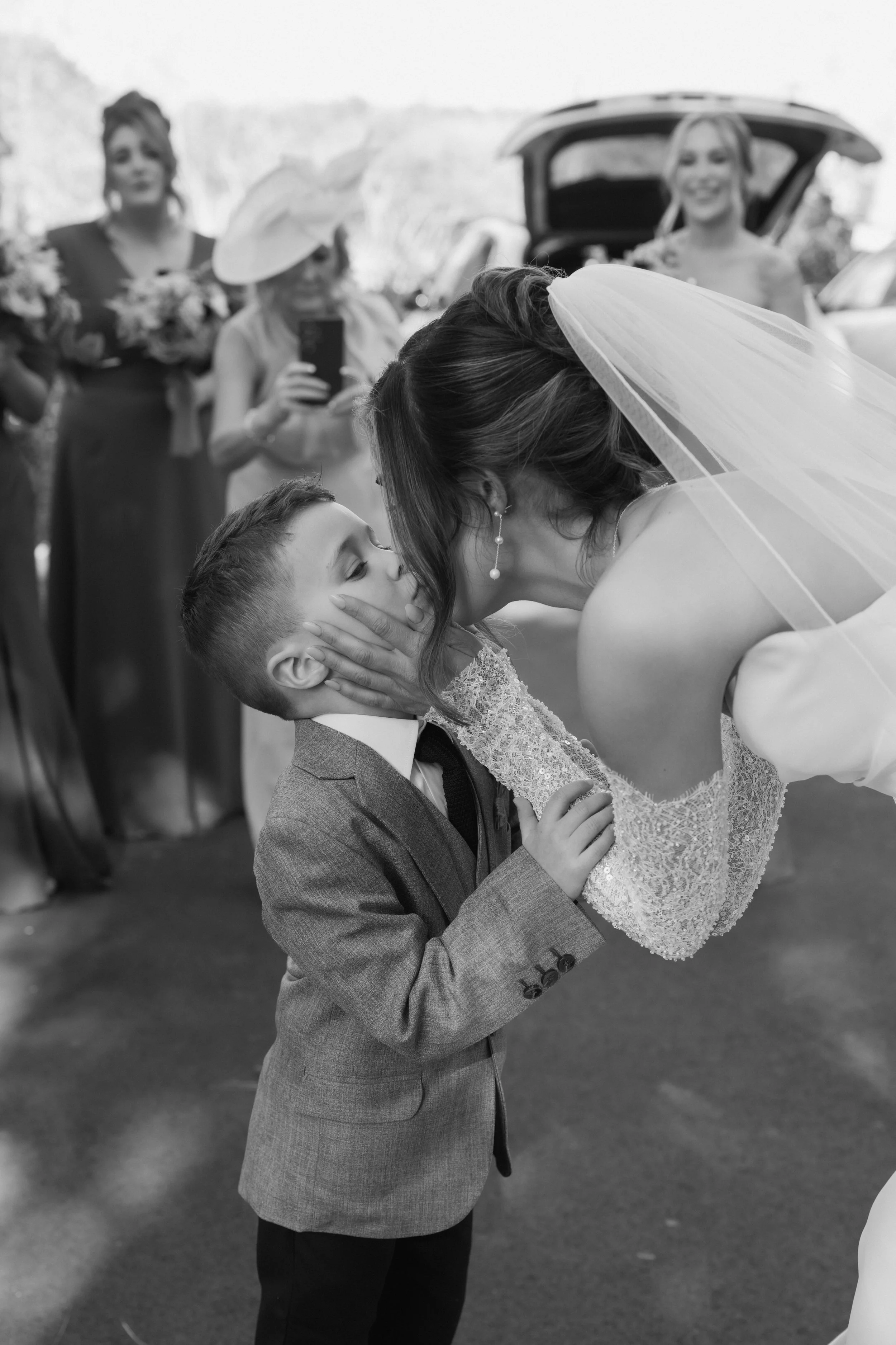 A bride leaning down to kiss a young boy in a suit at a wedding, with bridesmaids and guests in the background.