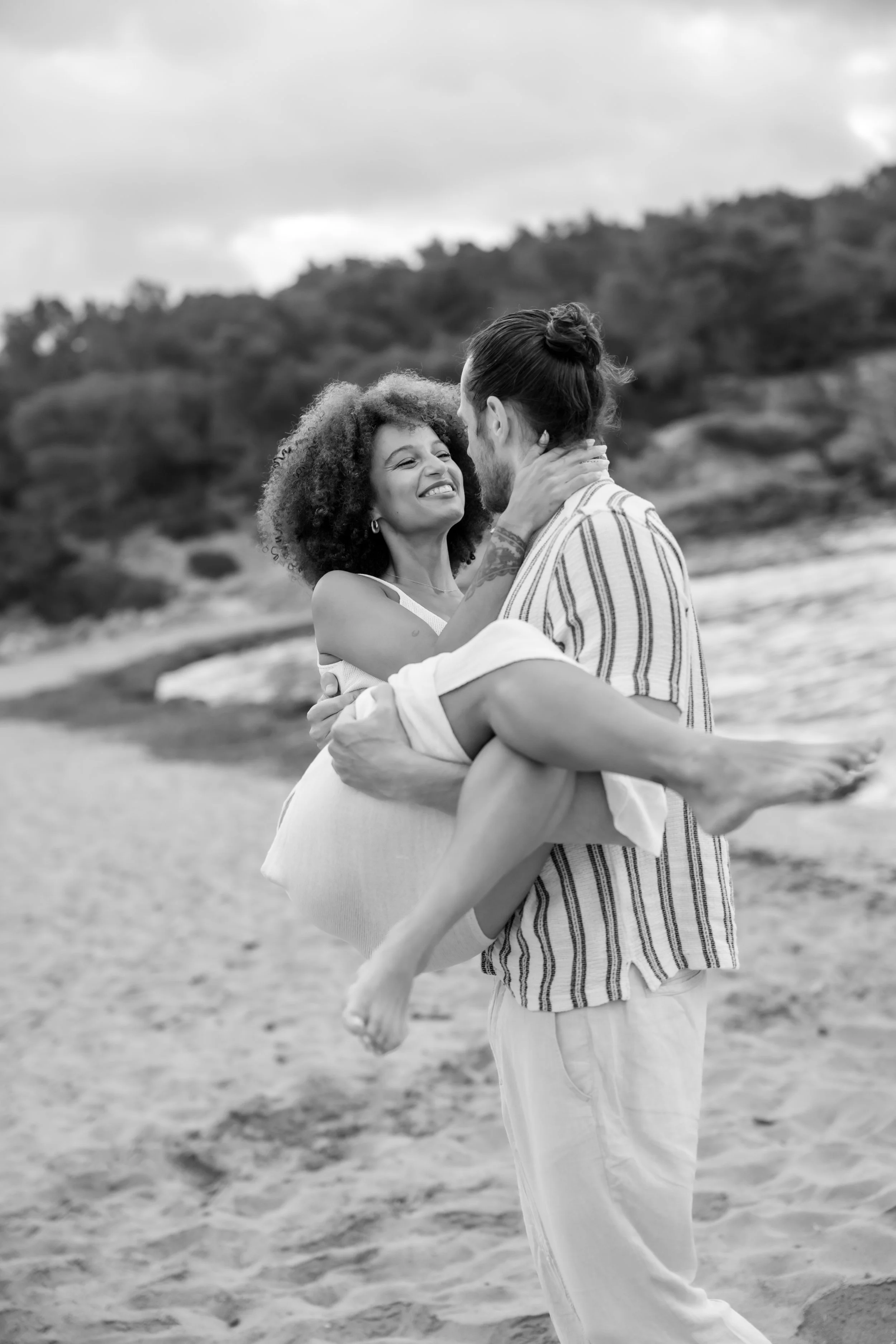 Man carrying woman in his arms on a beach with hilly landscape in the background, black and white photo.