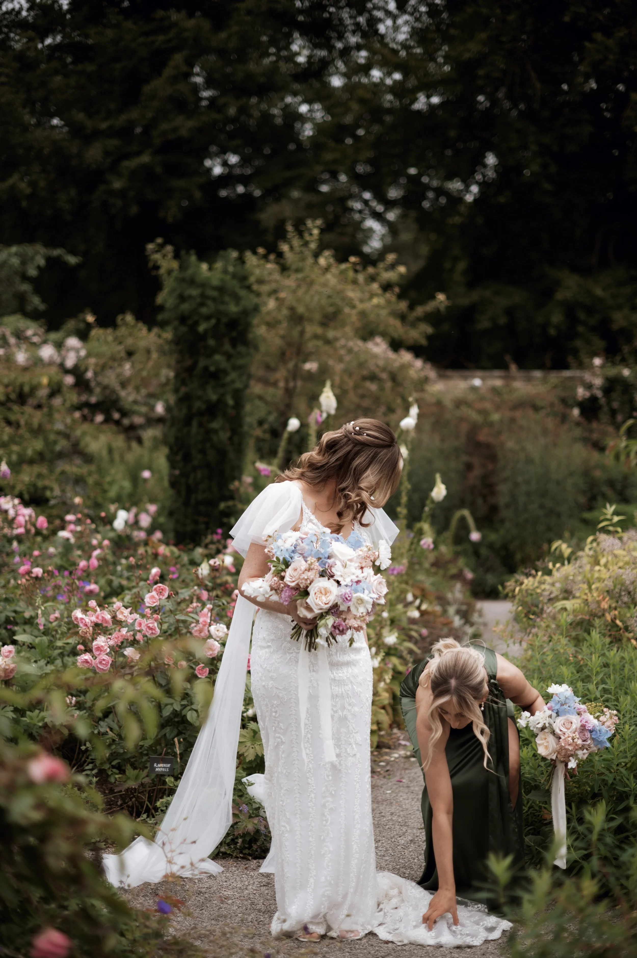 Bride holding pastel bouquet assisted by bridesmaid in a garden at Middleton Lodge 
