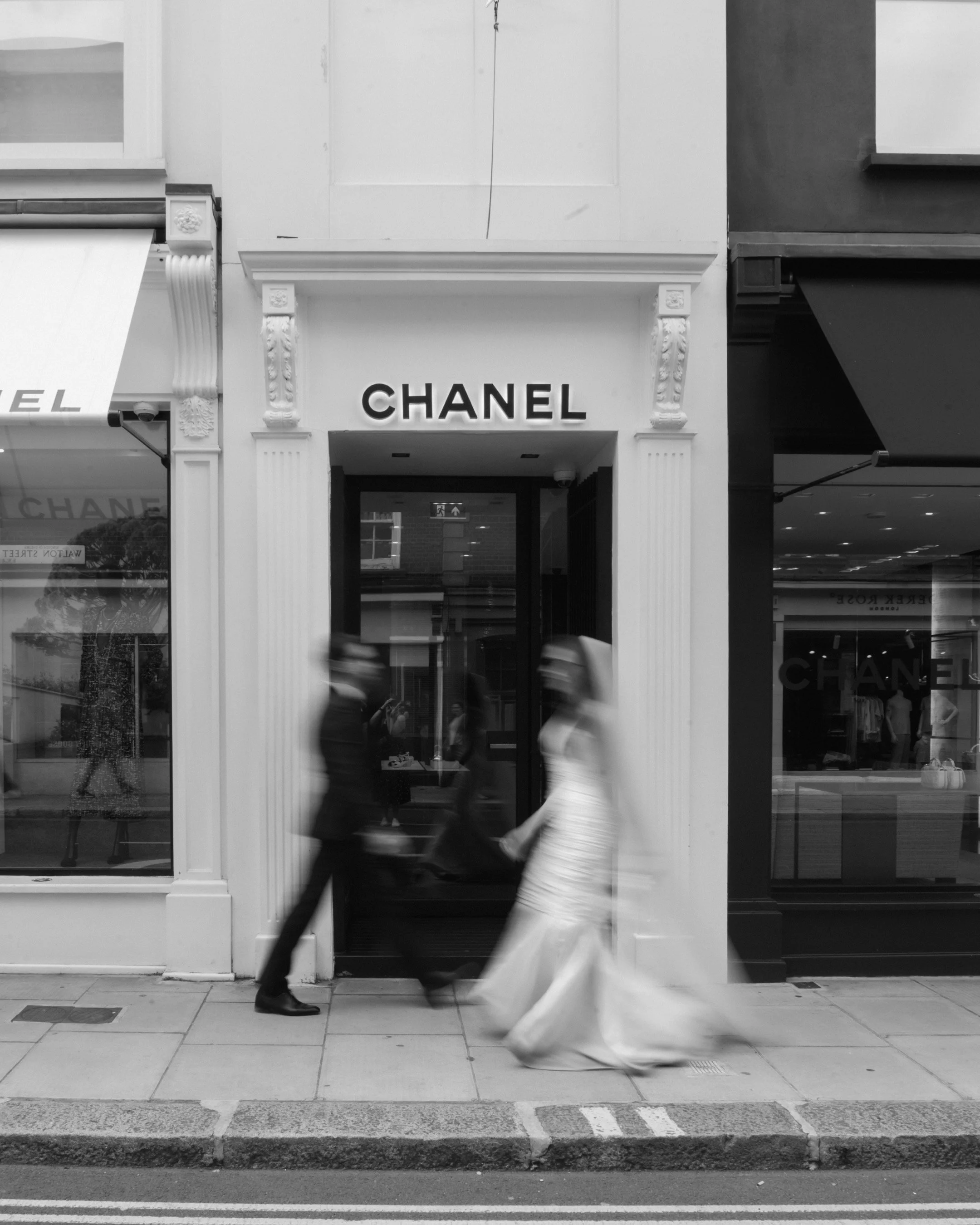 Black and white photo of a Chanel store with a bride and groom walking past in wedding attire, both appearing blurred due to motion.