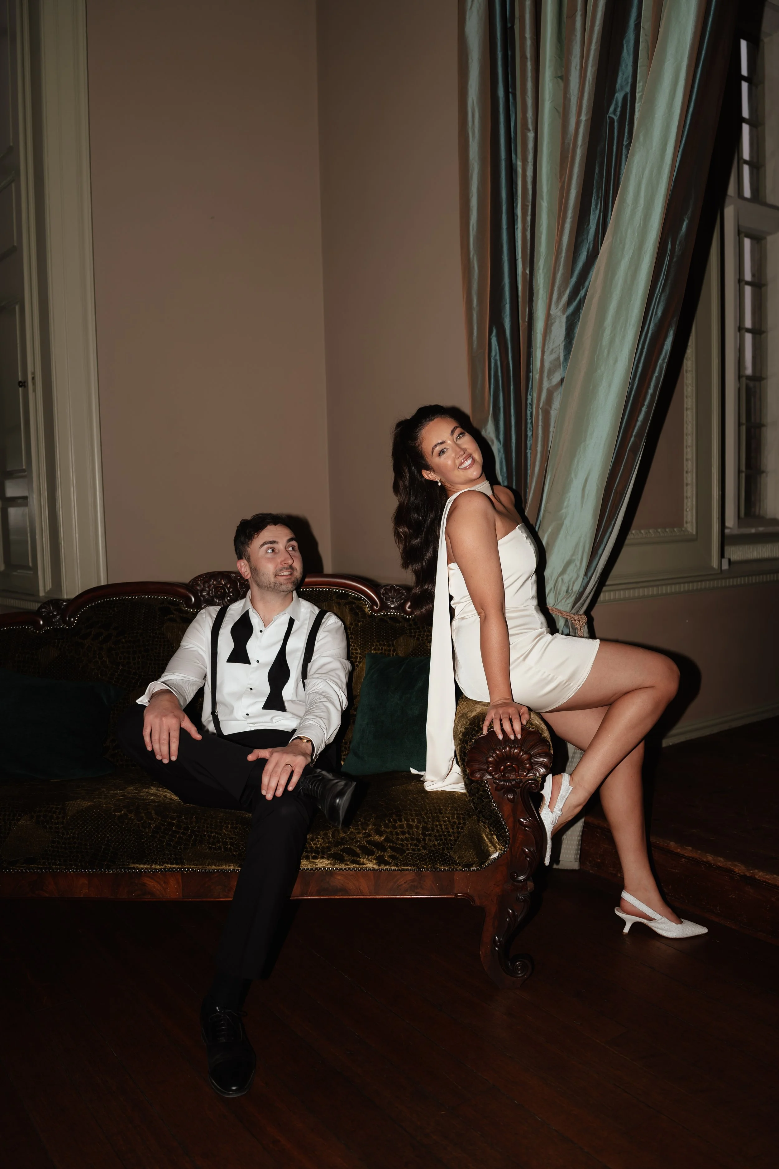Bride in white dress sitting on vintage sofa armrest, groom in tuxedo seated at Howsham Hall 