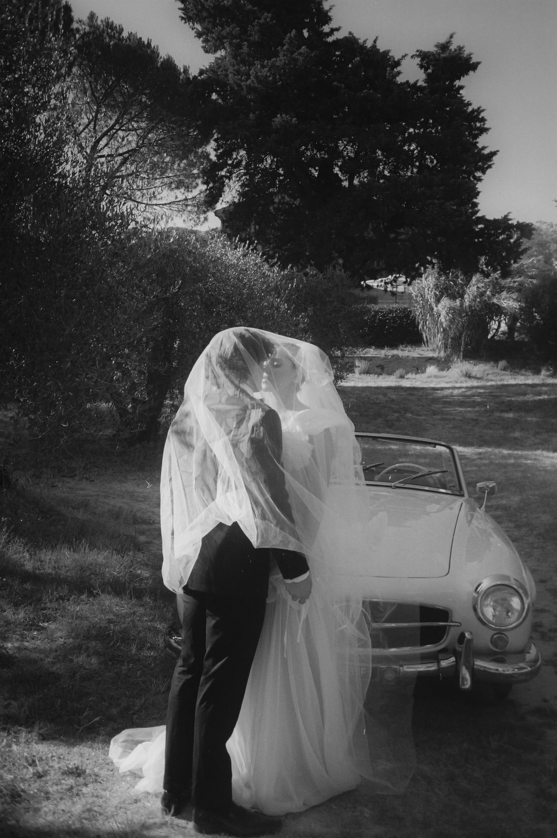 A bride and groom standing outside near a vintage convertible car, with the bride wearing a veil and wedding gown, and the groom in a suit, during daytime.