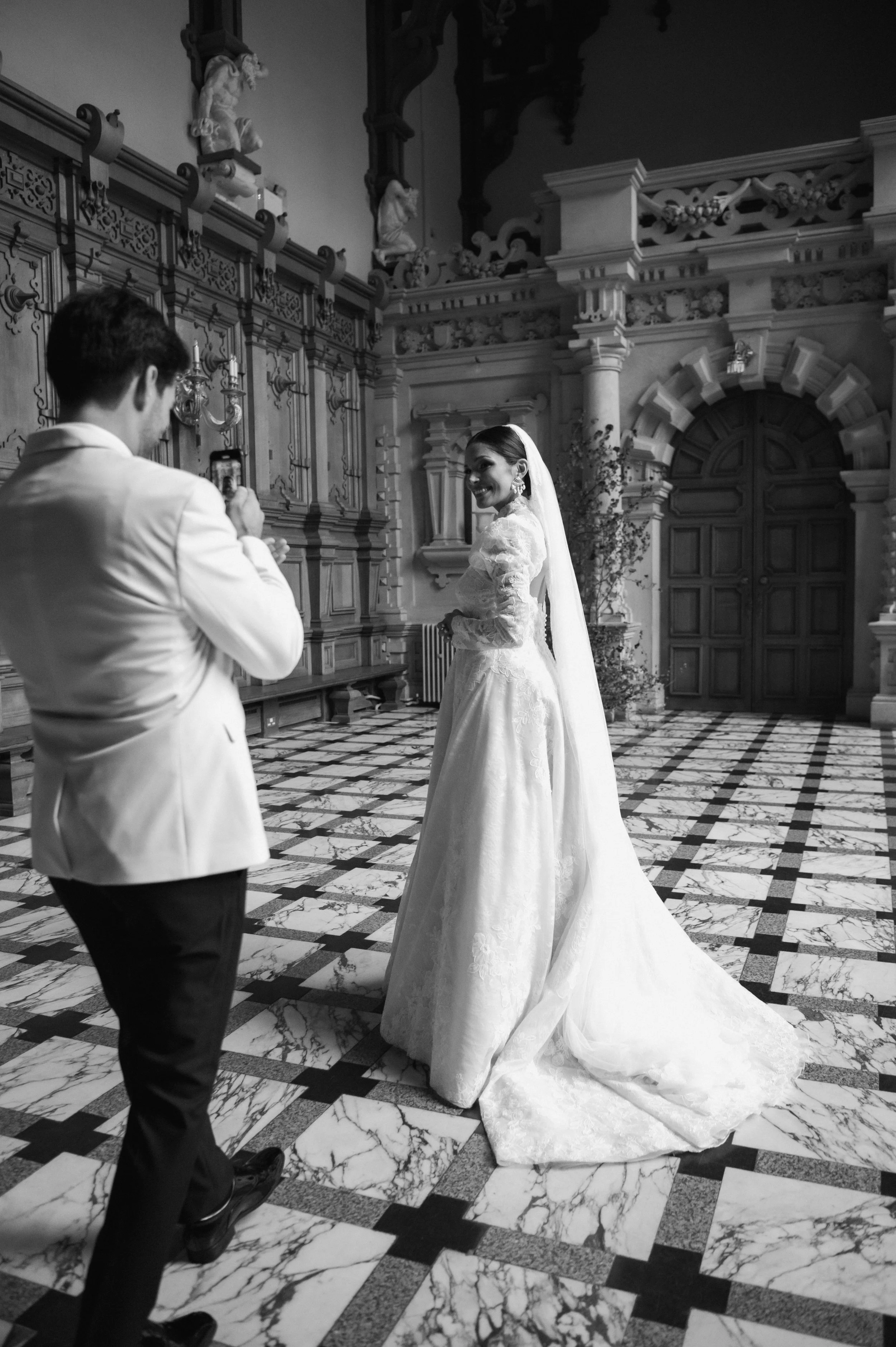 Bride smiling for photo while groom photographs her indoors with ornate decor at Harlaxton Manor