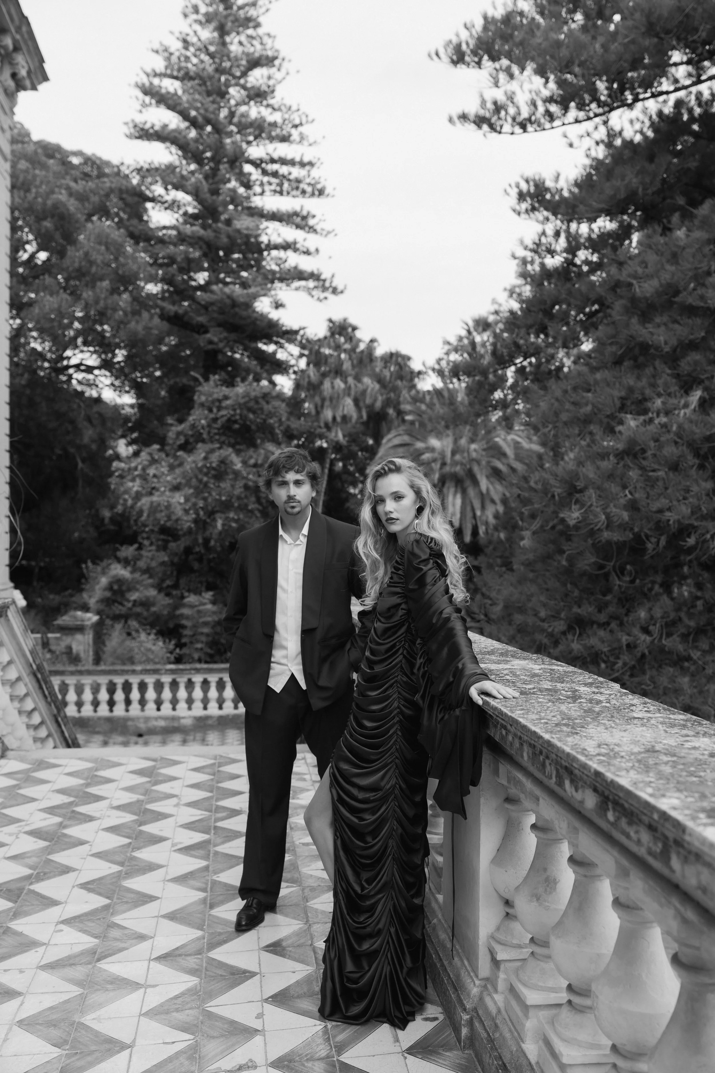 A black-and-white photo of a man and woman on a balcony with ornate balustrades, surrounded by tall trees in the background. The woman is leaning on the railing, dressed in a ruched, flowing gown, and the man stands slightly behind her, wearing a tux