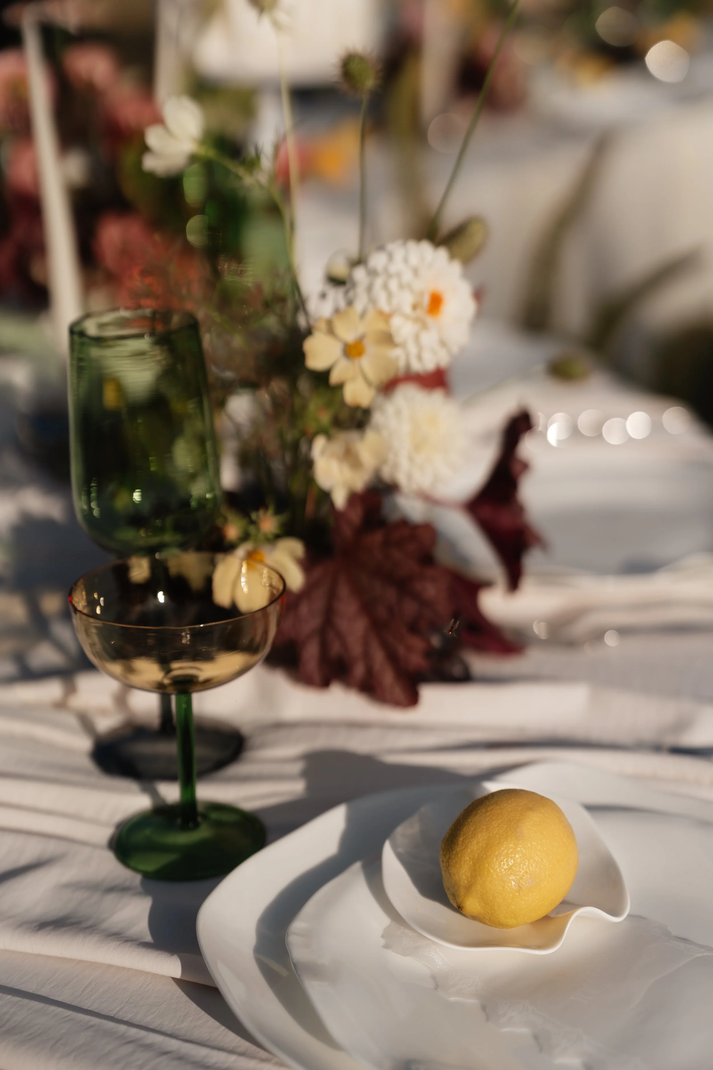 A lemon on a white plate with a white napkin underneath, set on a table alongside empty glasses and a floral centerpiece in natural sunlight.