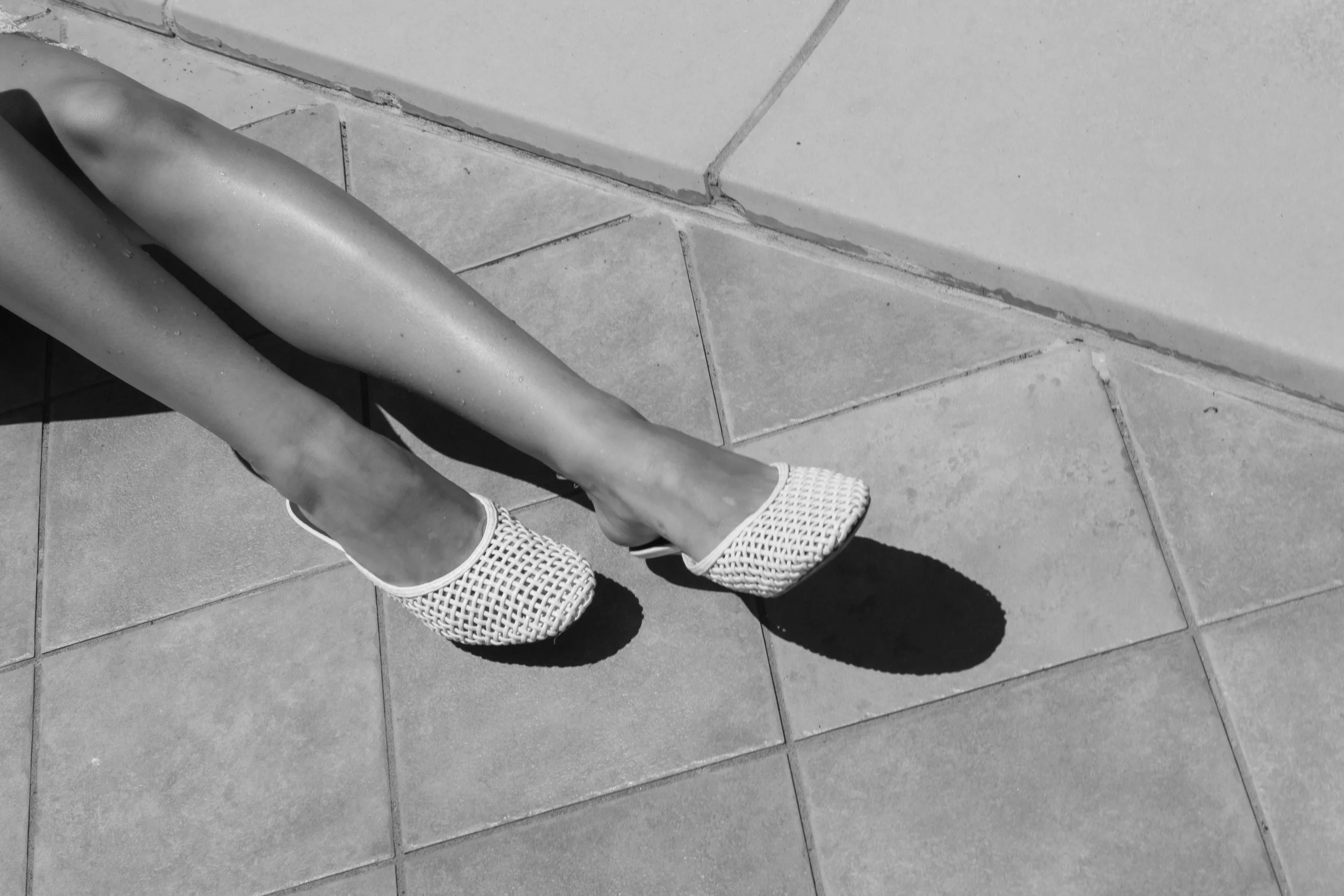 Black and white photo of a person's legs and feet wearing woven style shoes, sitting on a tiled surface with their legs crossed. Their shadow is visible on the ground.