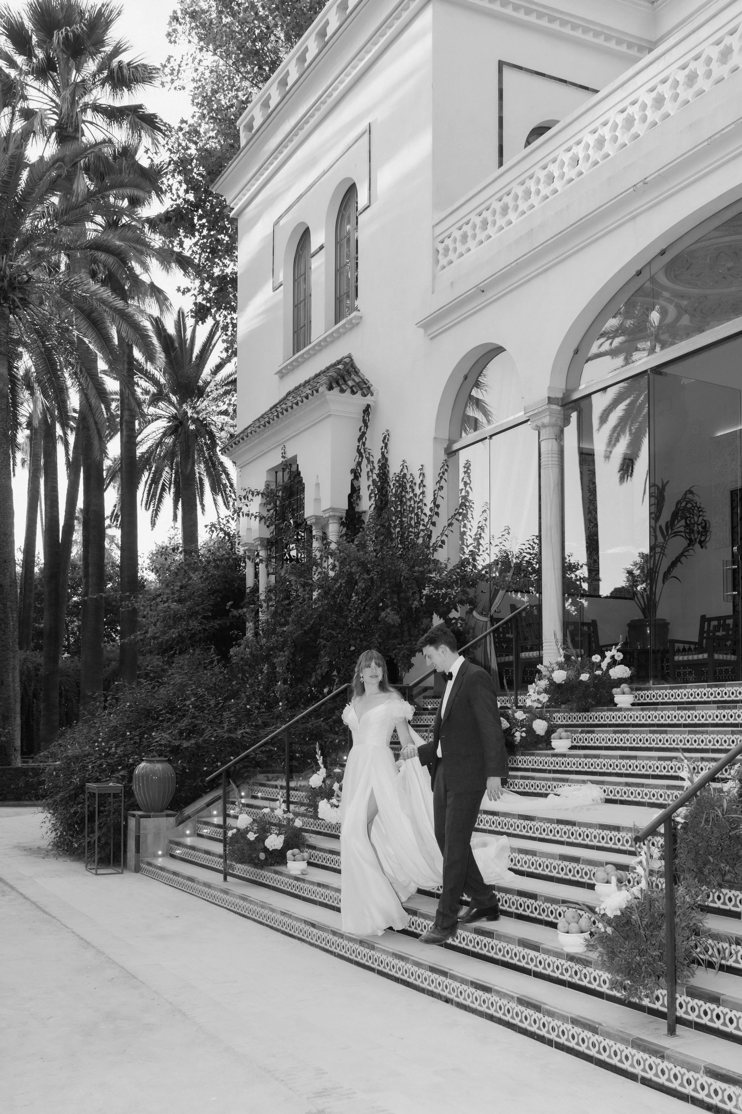 A bride and groom descending outdoor stairs decorated with flowers and fruit outside a large white building with palm trees nearby at Villa Luisa