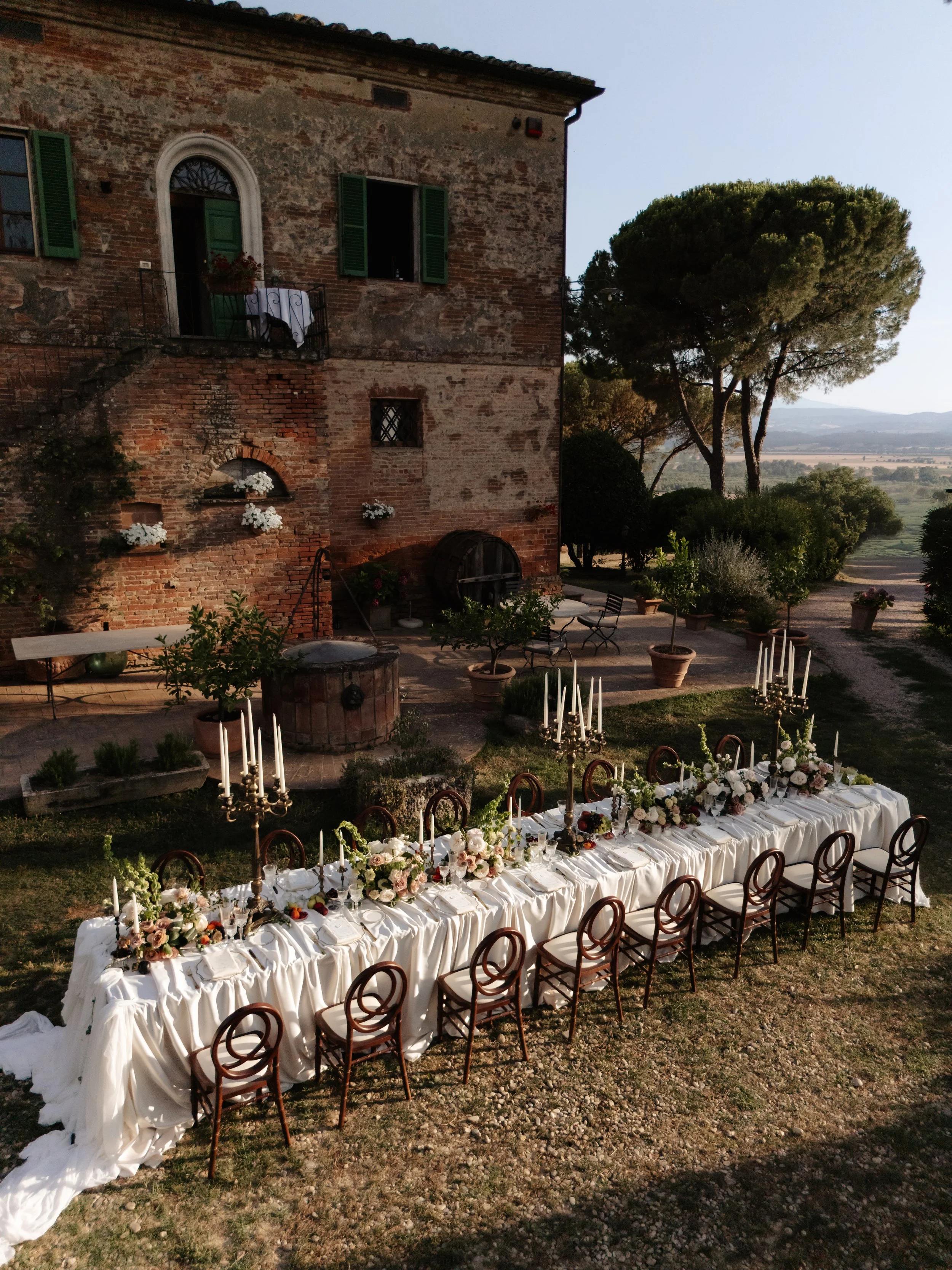 A long dining table with white tablecloths, floral centerpieces, and candelabras set outdoors near a brick building and trees.