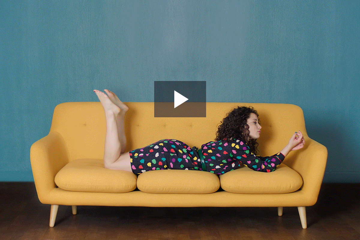 A woman with curly hair lying on her stomach on a yellow couch with beige cushions, wearing a colorful polka dot dress, against a teal wall background.