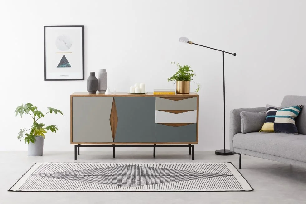 Modern living room with a wooden sideboard, potted plants, wall art, floor lamp, gray sofa, and a striped cushion, all arranged on a striped rug against a white wall.