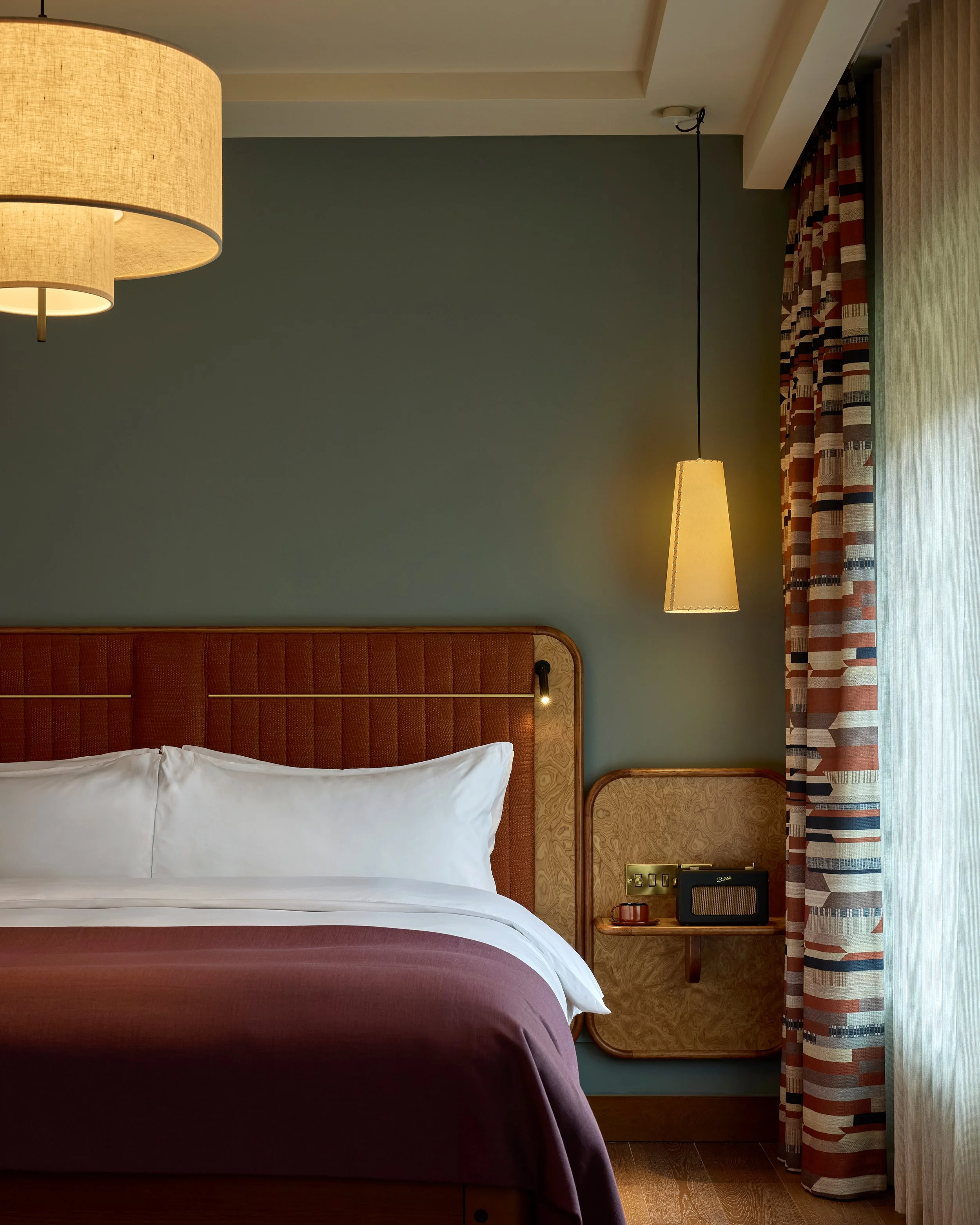 A hotel room with a bed, white pillows, and a maroon bedspread. A large window with patterned curtains and a small bedside table holding a vintage radio, a cup, and some electrical outlets. Two hanging lamps, one large ceiling light and one narrow pe