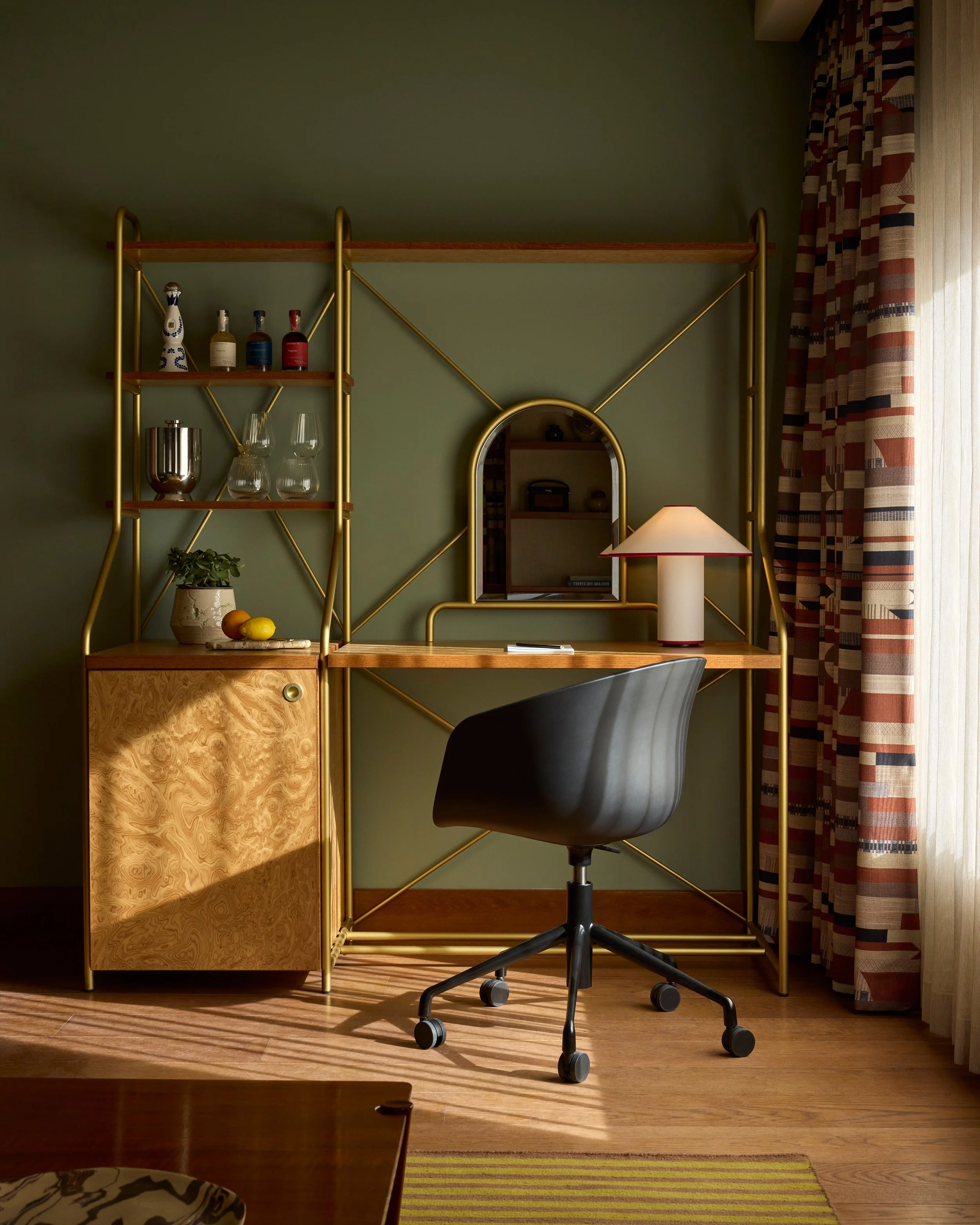 A home office desk with a black swivel chair in front of a green wall, gold metal frame shelving, and a small mirror, desk lamp, and decorative items.
