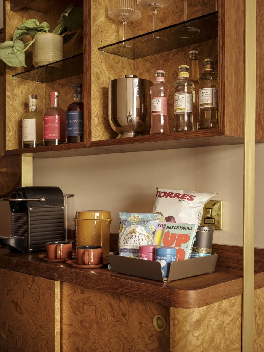 Wooden shelves with bottles of alcohol, glasses, a potted plant, a coffee machine, cups, snacks, and a storage bin.