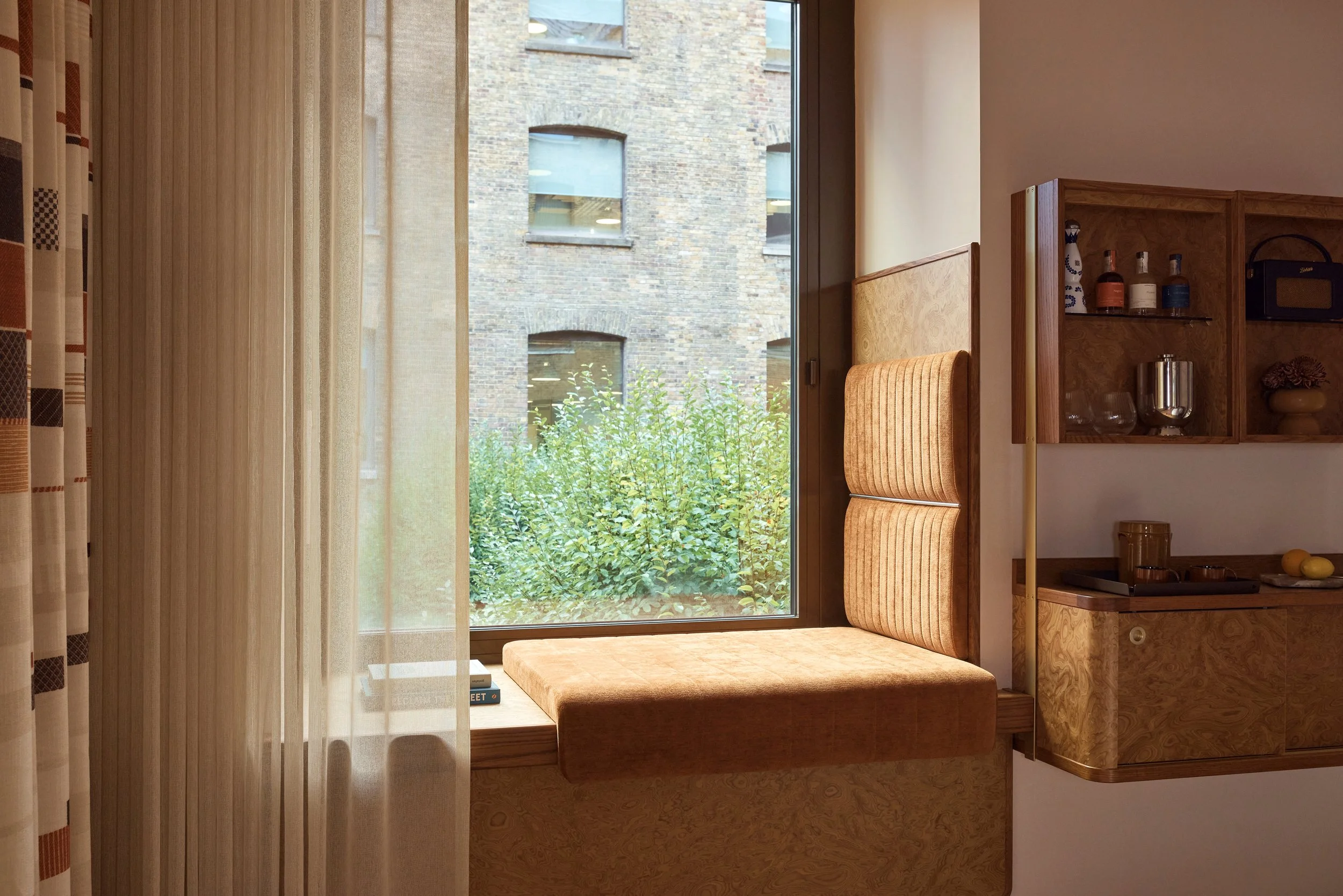 A cozy window seat with cushioned backrest and a view of a brick building outside, flanked by curtains on the left and a wooden cabinet with bottles and decorative items on the right.