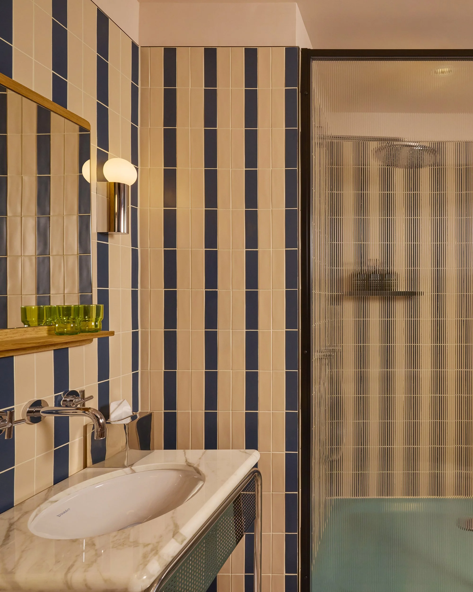 Devonshire Square hotel bathroom with pink and navy striped tiled walls, a sink with a marble countertop, a mirror, a wall-mounted light fixture, a small shelf with green glasses, and a glass shower door with textured glass design.