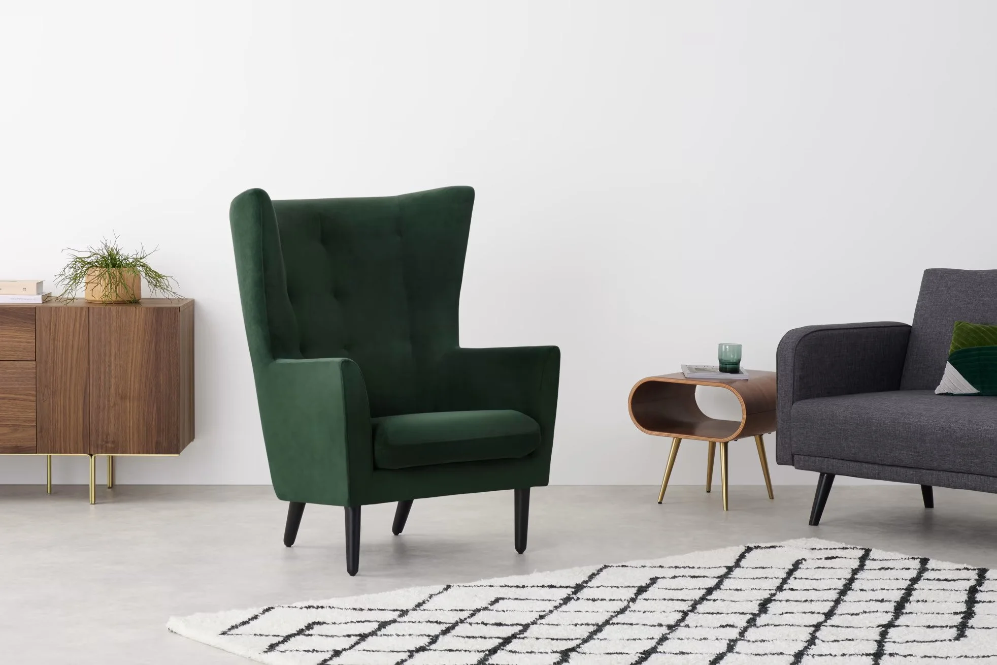 Modern living room with green wingback chair, gray sofa with decorative pillows, wooden side table with books and plant, and a wooden cabinet against a plain white wall, with a black and white patterned rug on the floor.