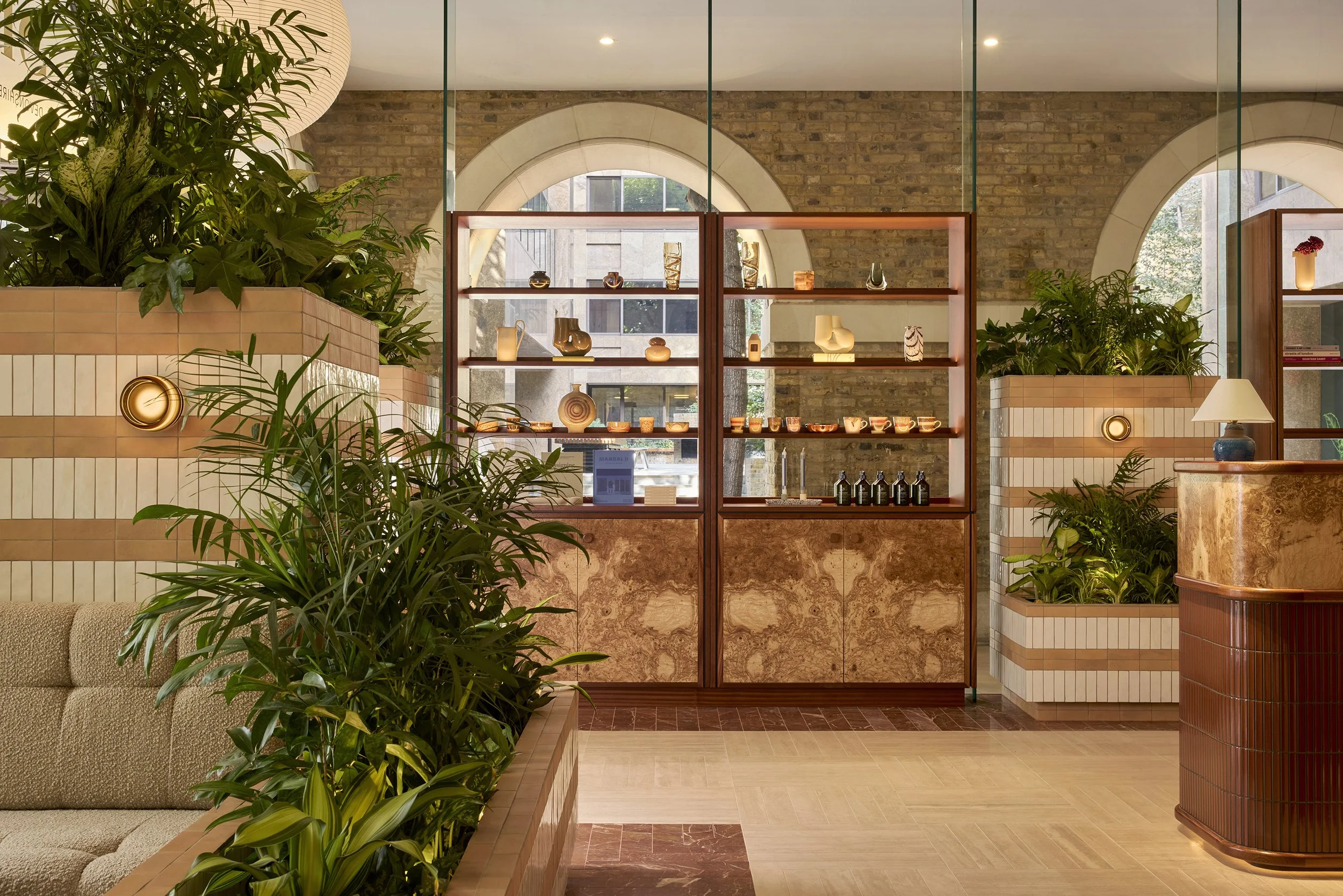 Interior of a modern boutique hotel lobby with wooden display shelves, potted plants, a marble countertop, and decorative glassware and ceramics.