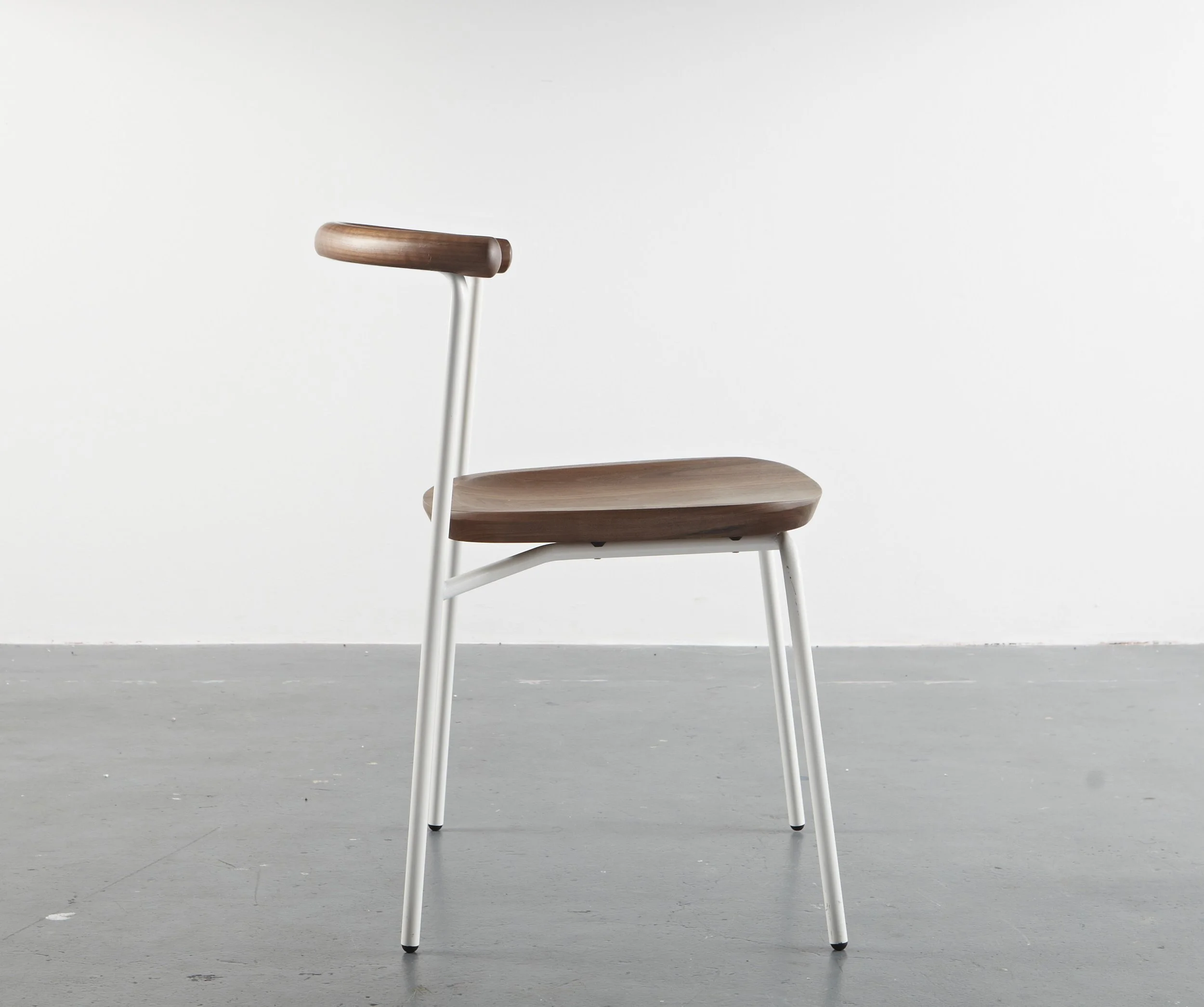 Modern chair with wooden seat and backrest, white metal frame, placed on gray concrete floor against plain white wall.