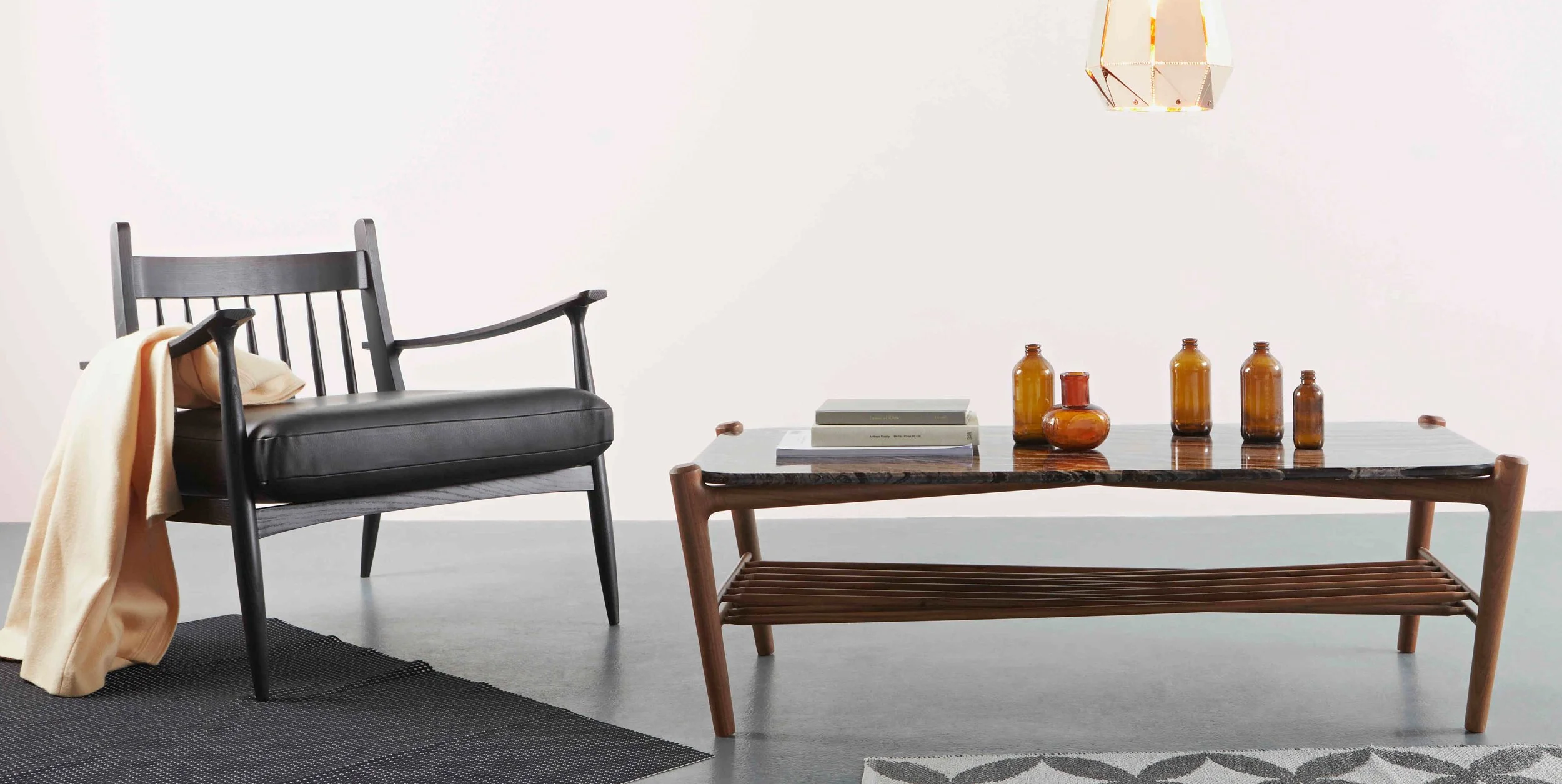 Modern living room with a black armchair, beige throw blanket, wooden coffee table with amber glass vases, books, and a wall-mounted geometric lamp.
