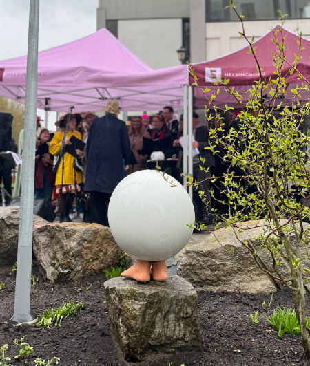 En utomhusscen med ett fåtal pinkvantäckta tält och en grupp människor under ett av tälten. Det finns en konstnärlig skulptur av ett ägg på en sten i förgrunden. Skulpturen har två små händer som stöder den.