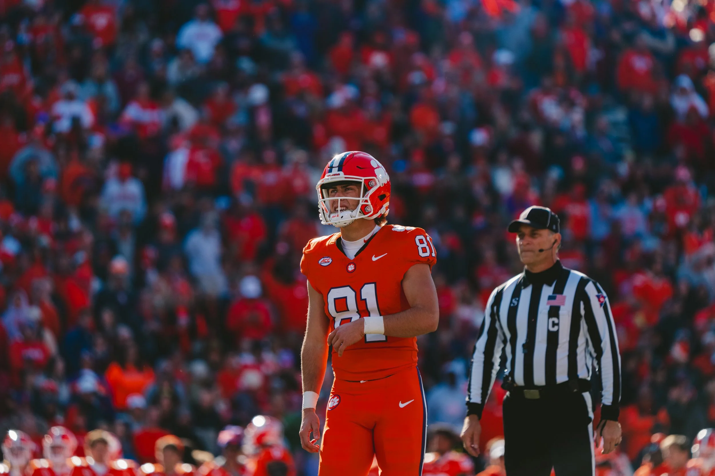 A football player in an orange uniform with a helmet stands on the field with an referee nearby, and a crowd of spectators in red in the background.