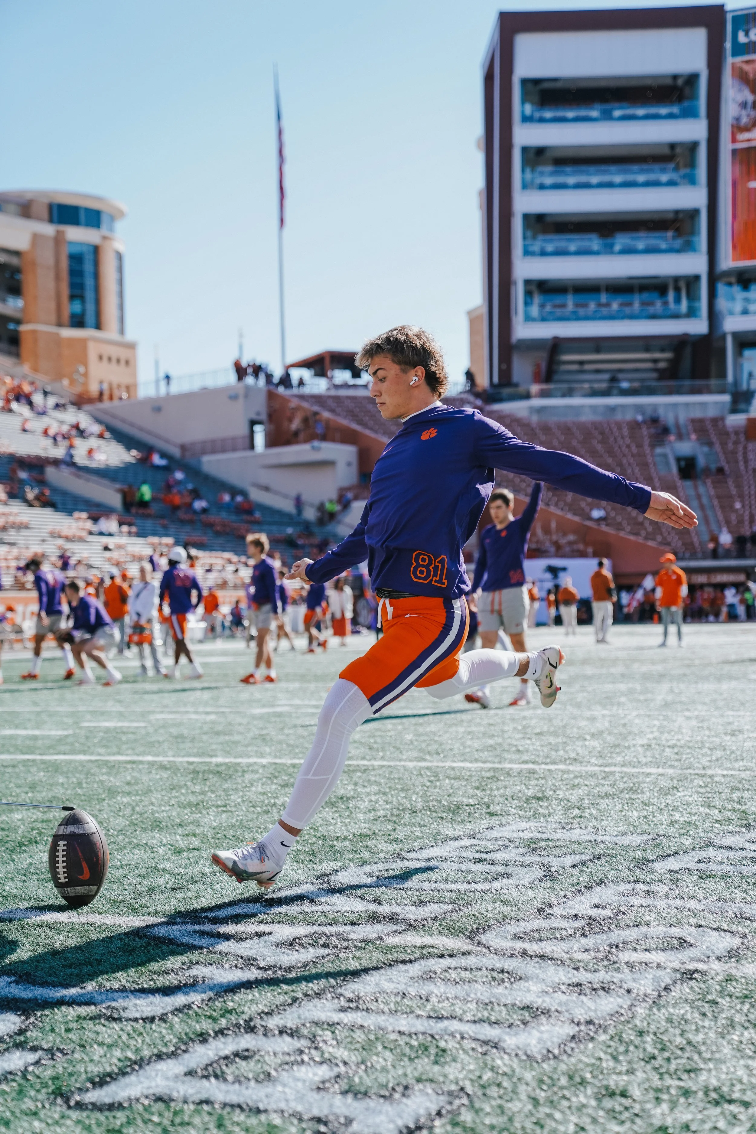 A football player in a blue jersey with the number 81 and orange shorts kicks a football on a stadium field, with other players and fans in the background.