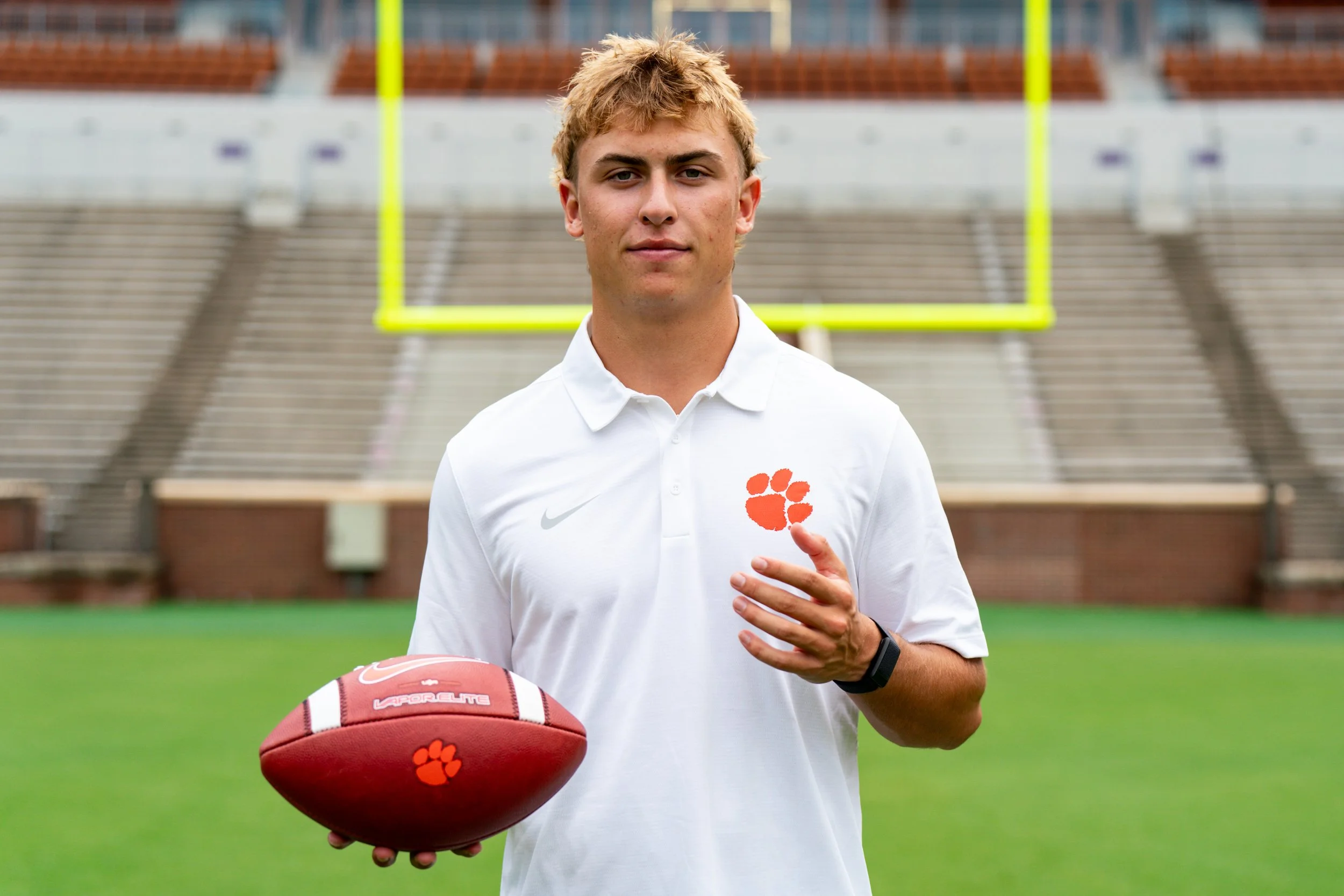 Young male athlete standing on a football field holding a football, wearing a white Nike polo shirt with an orange paw print logo, standing in front of a goalpost in a stadium.