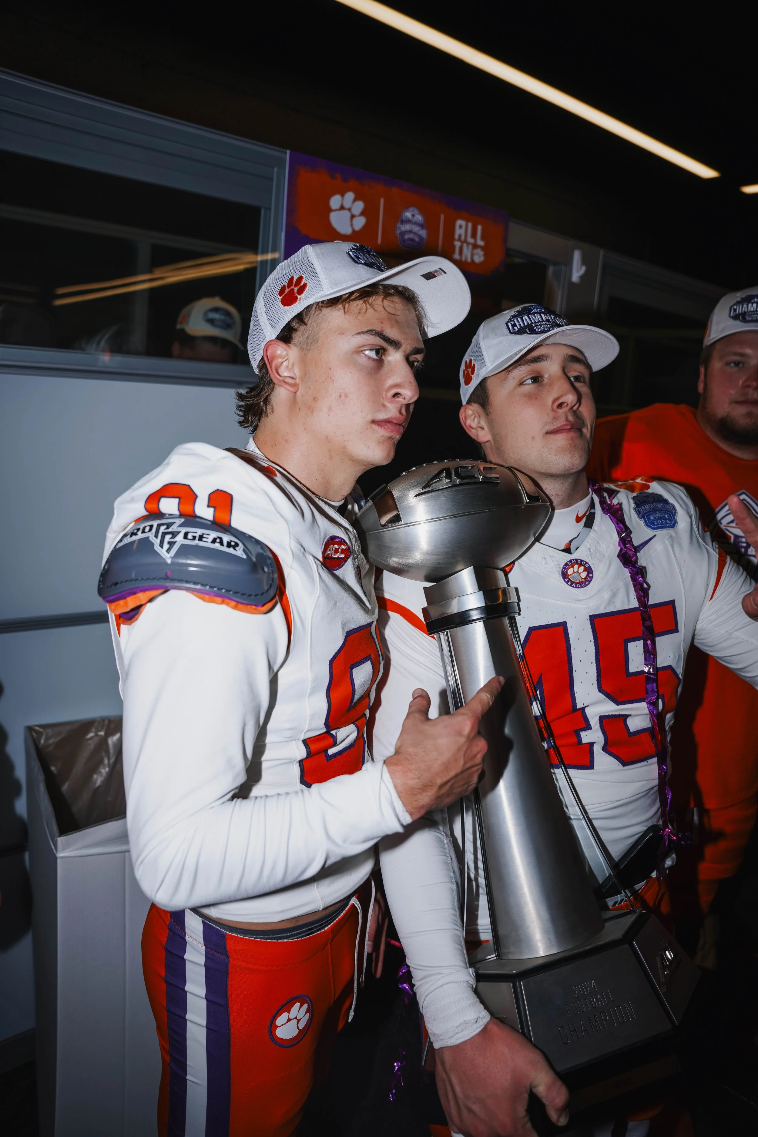 Two young football players in white and orange uniforms holding a trophy; one is pointing at the trophy. They are in a locker room or team area, wearing white caps with a paw print logo, with a background featuring a sign that says 'All In' and a ref